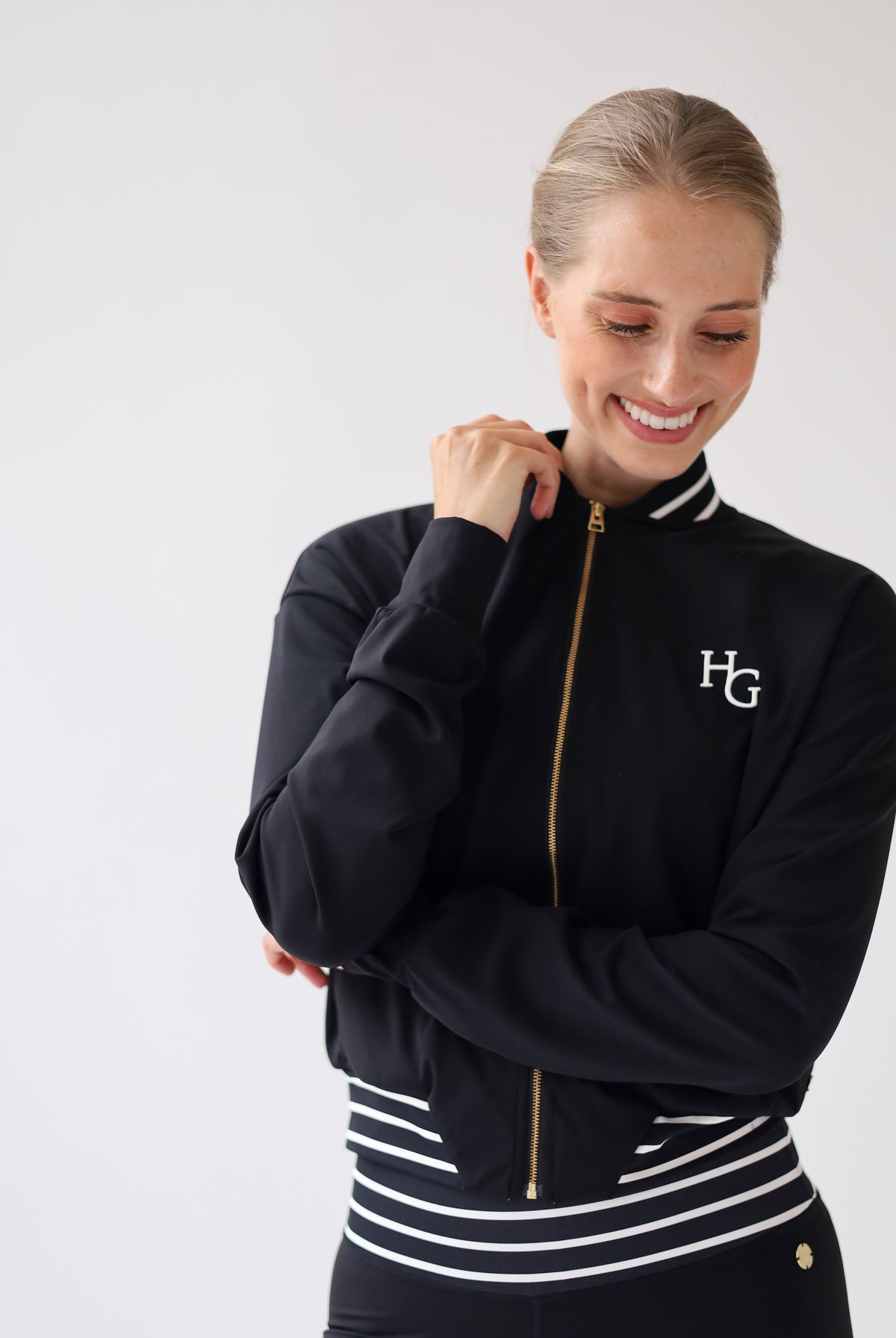 A woman wearing a black zip-up athletic jacket with white stripes on the collar and hem, gold zipper details, and a white 'HG' logo on the left chest, posed against a light gray studio background.