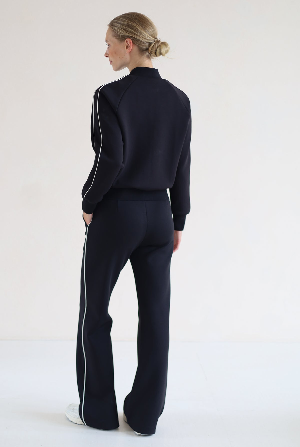 A woman stands with her back to the camera wearing a matching black set with white side stripes, consisting of a cropped mock-neck jacket and wide-leg pants, against a plain white studio background.