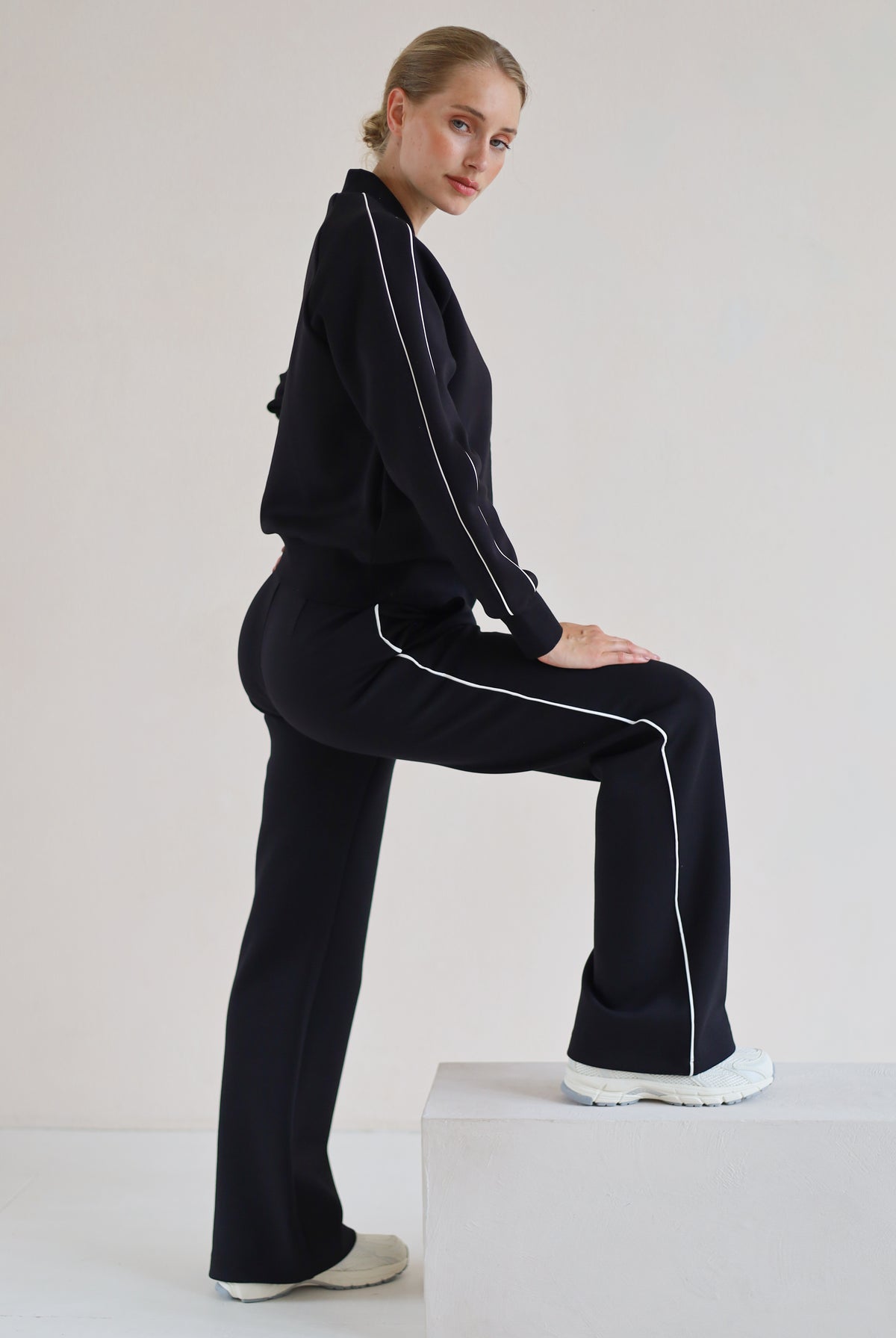 A woman models a matching black set with white piping details, posing on a white block against a plain light background.
