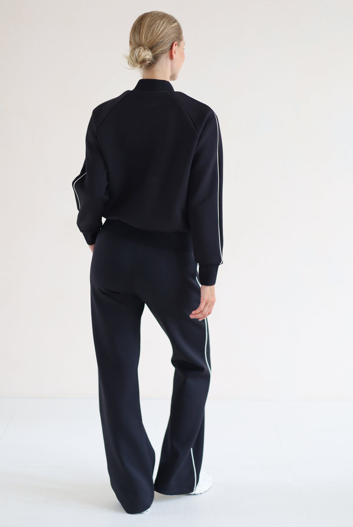 A woman viewed from behind wearing a blackbomber jacket with white side stripes and matching black wide-leg trousers with white side stripes, standing against a plain white studio background.