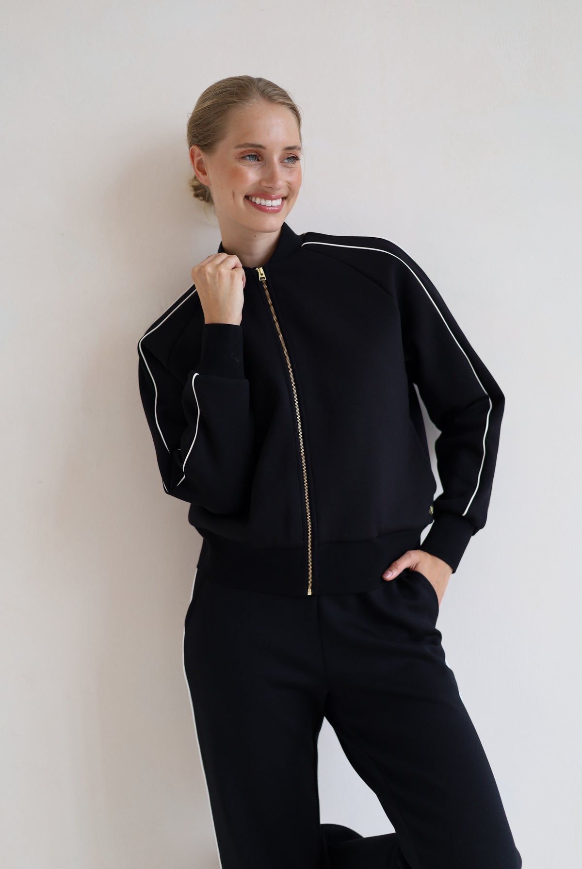 A woman wearing a black zip-up bomber jacket with white piping and a gold zipper, paired with black wide leg trouser, posing against a plain off-white wall.