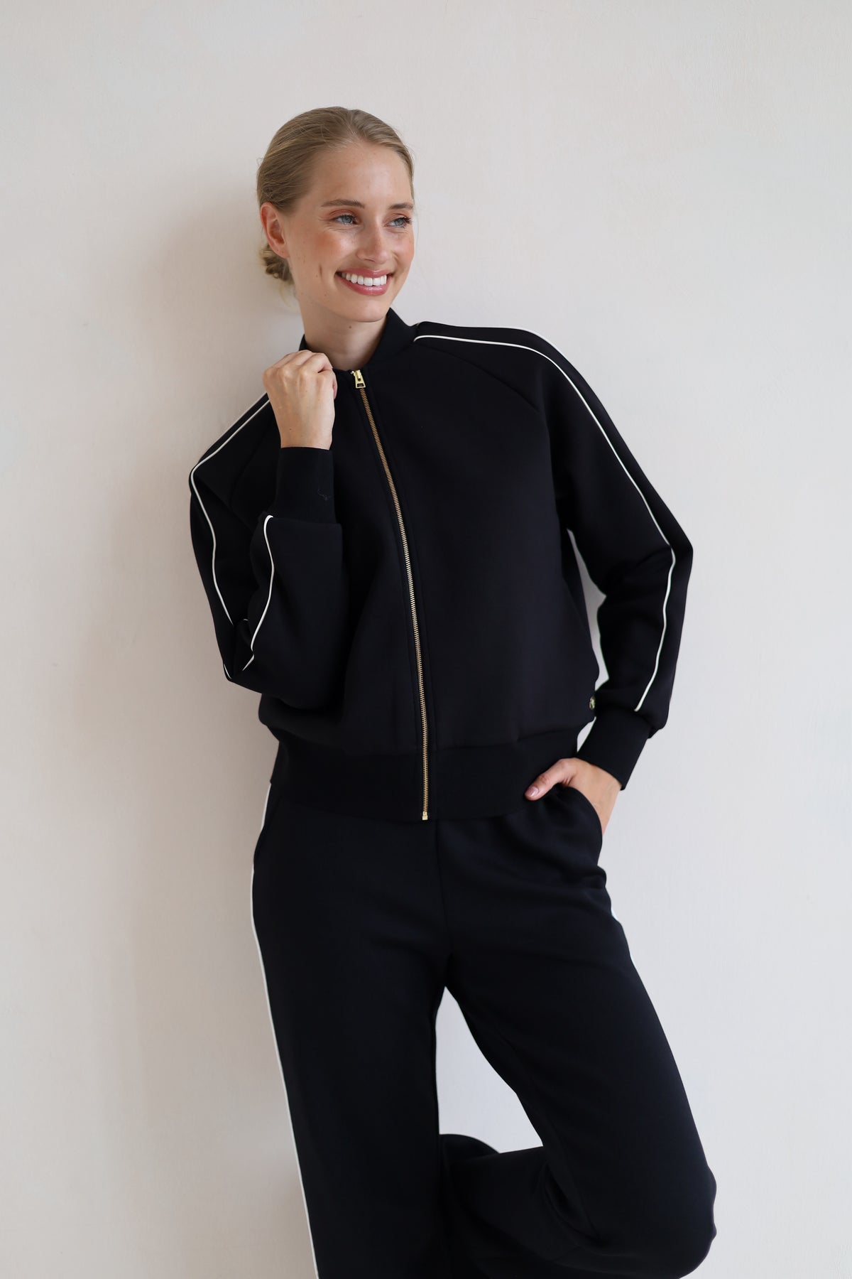 A woman wearing a black zip-up bomber jacket with white piping and a gold zipper, paired with black wide leg trouser, posing against a plain off-white wall.