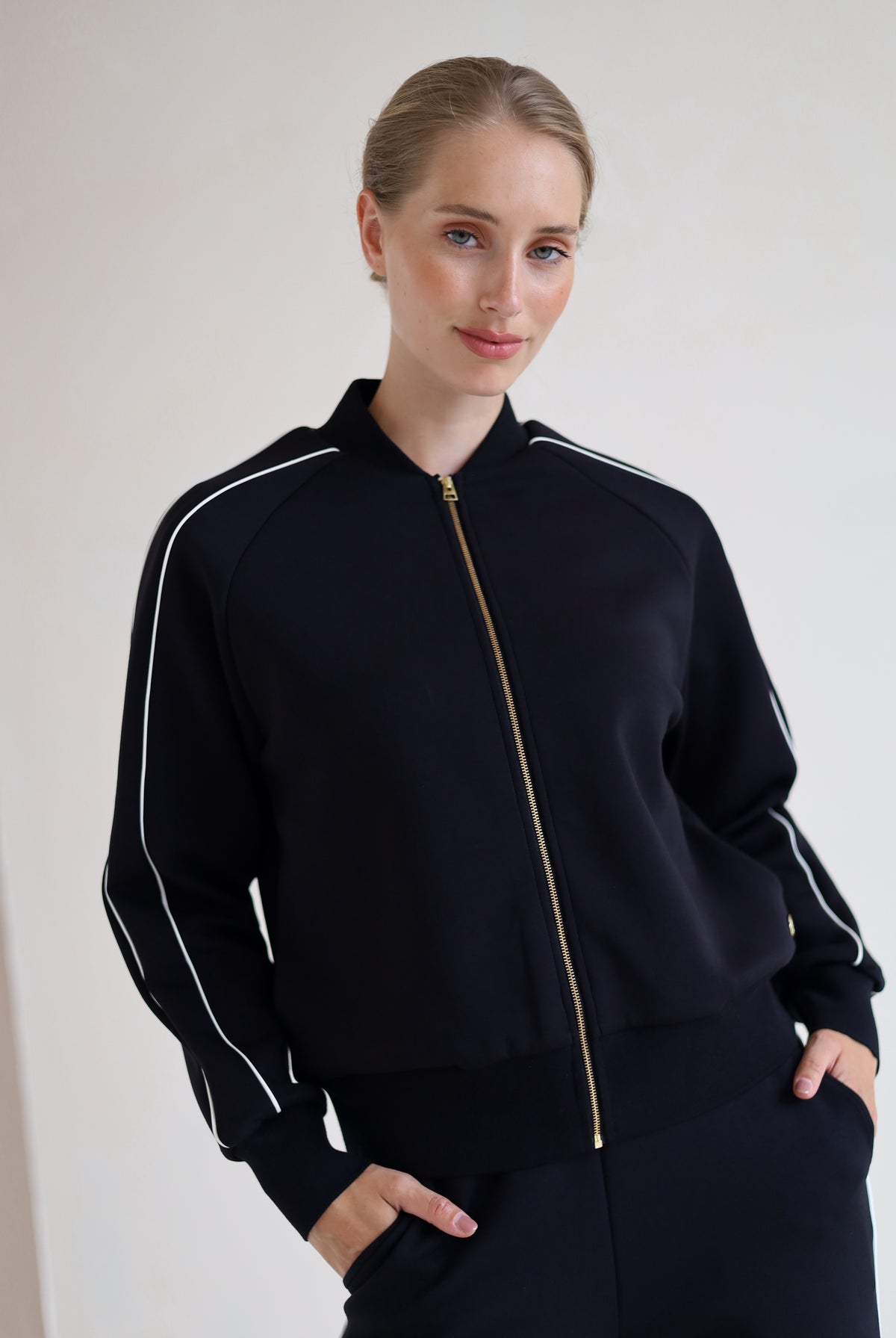 A woman wearing a black zip-up bomber jacket with white piping and a gold zipper, paired with black wide leg trouser, posing against a plain off-white wall.