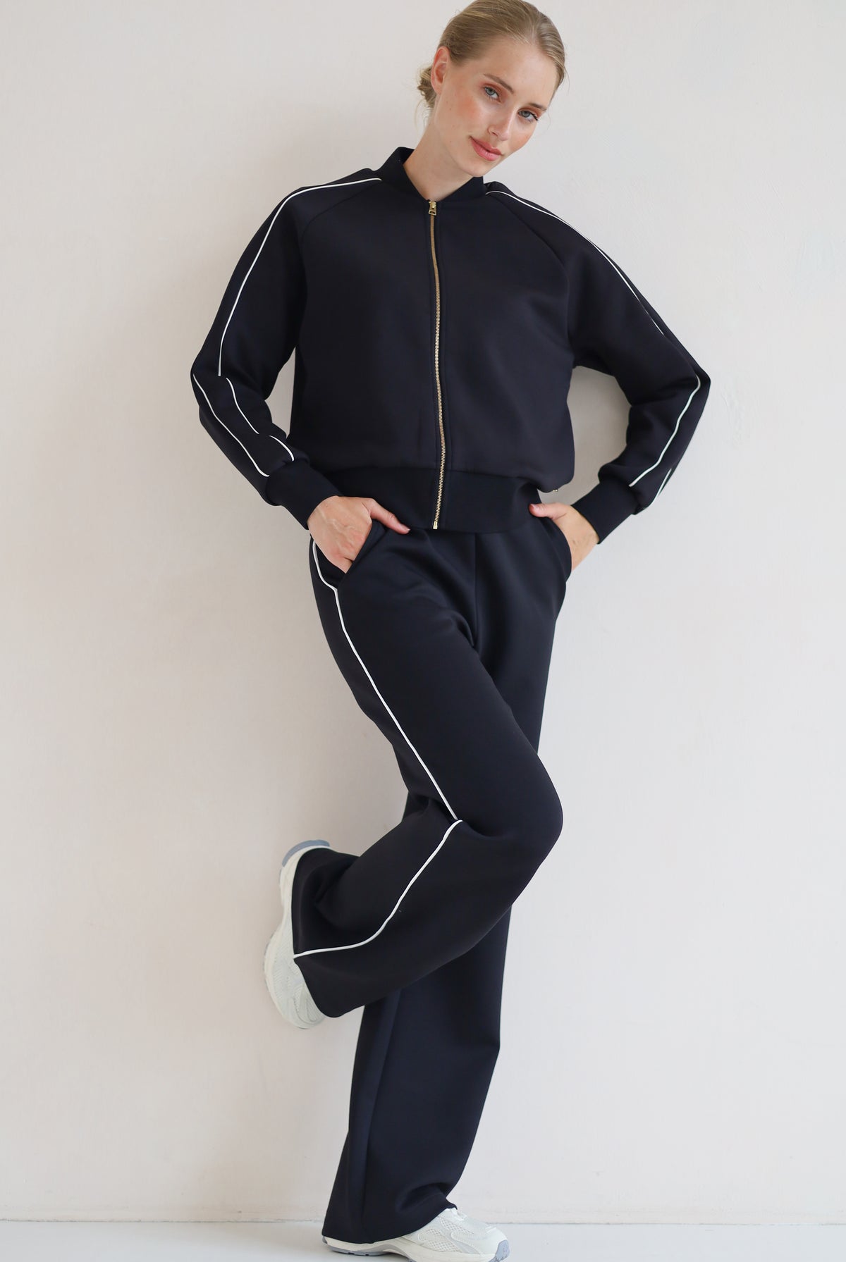 A woman modeling a matching black set with white piping details, featuring a zip-up bomber jacket and wide-leg pants, paired with white sneakers against a plain off-white background.