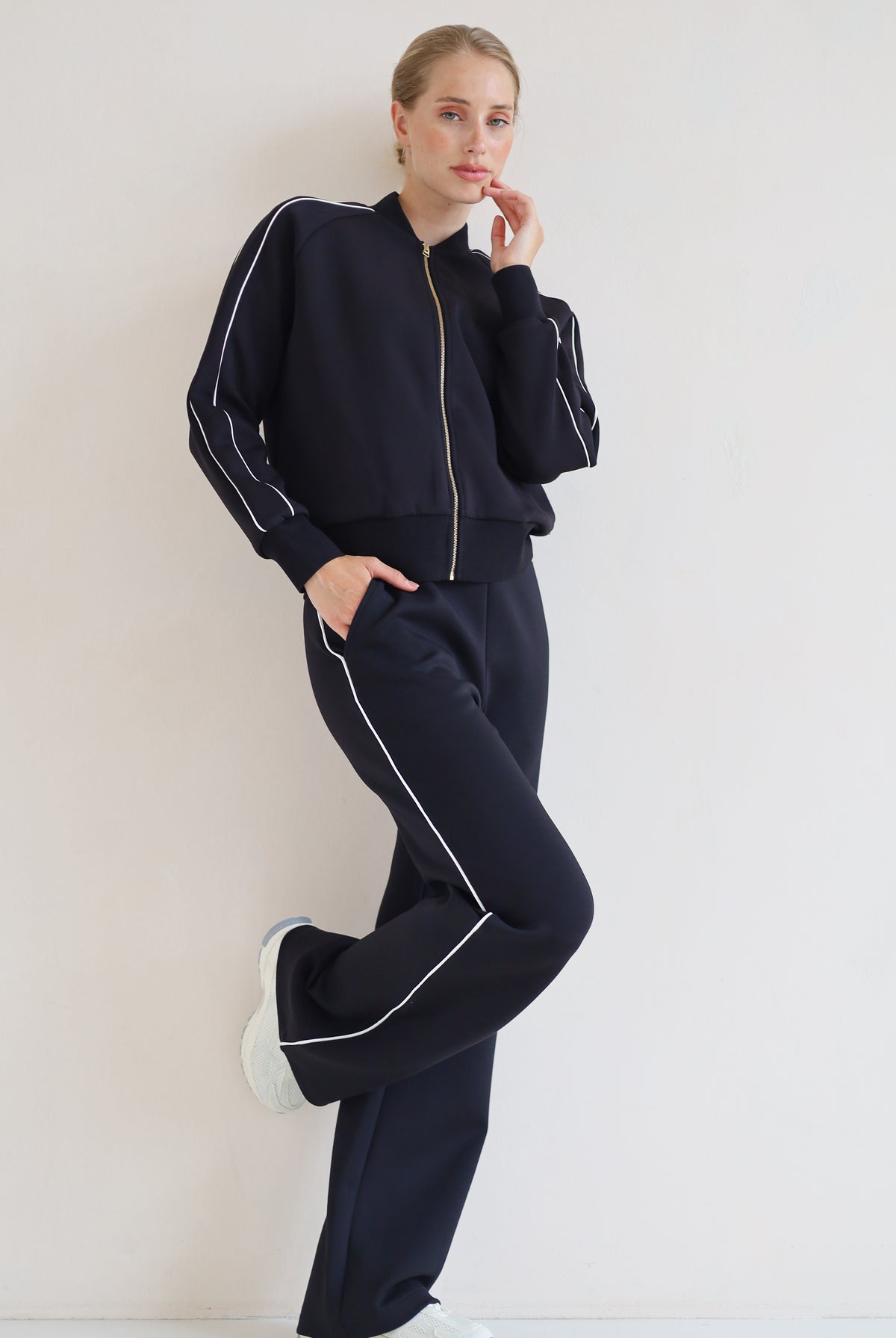 A woman modeling a matching black set with white piping details, featuring a zip-up bomber jacket and wide-leg pants, paired with white sneakers against a plain off-white background.