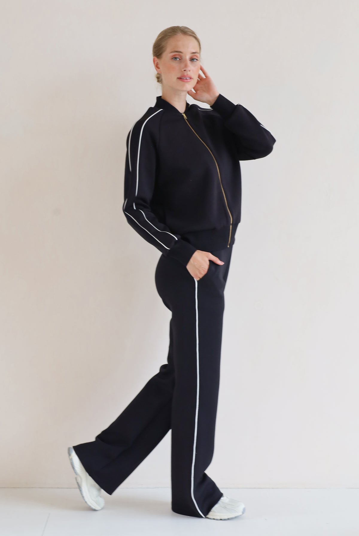A woman modeling a matching black set with white piping details, featuring a bomber jacket and wide-leg pants, set against a plain off-white studio background.