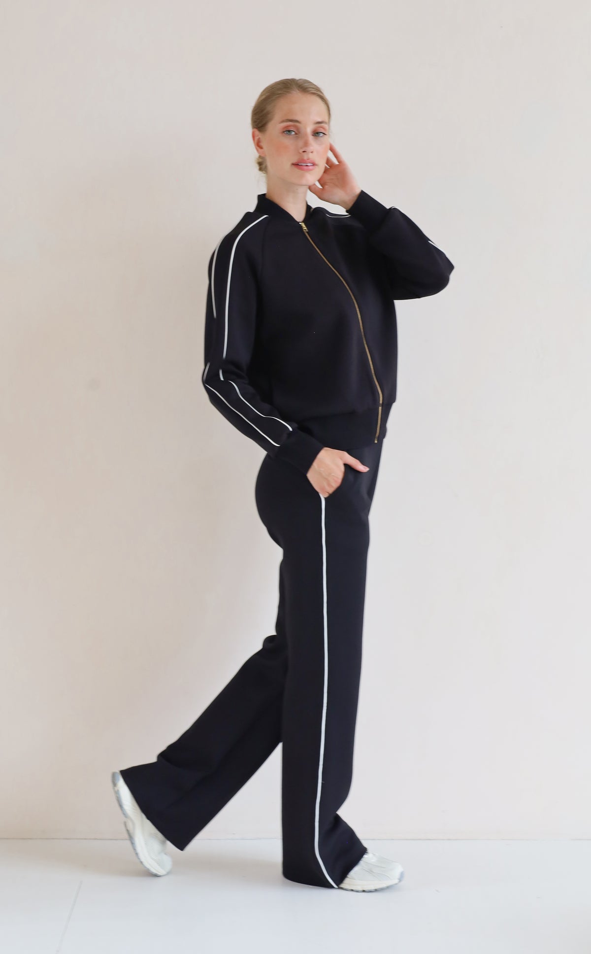 A woman modeling a matching black set with white piping details, featuring a bomber jacket and wide-leg pants, set against a plain off-white studio background.