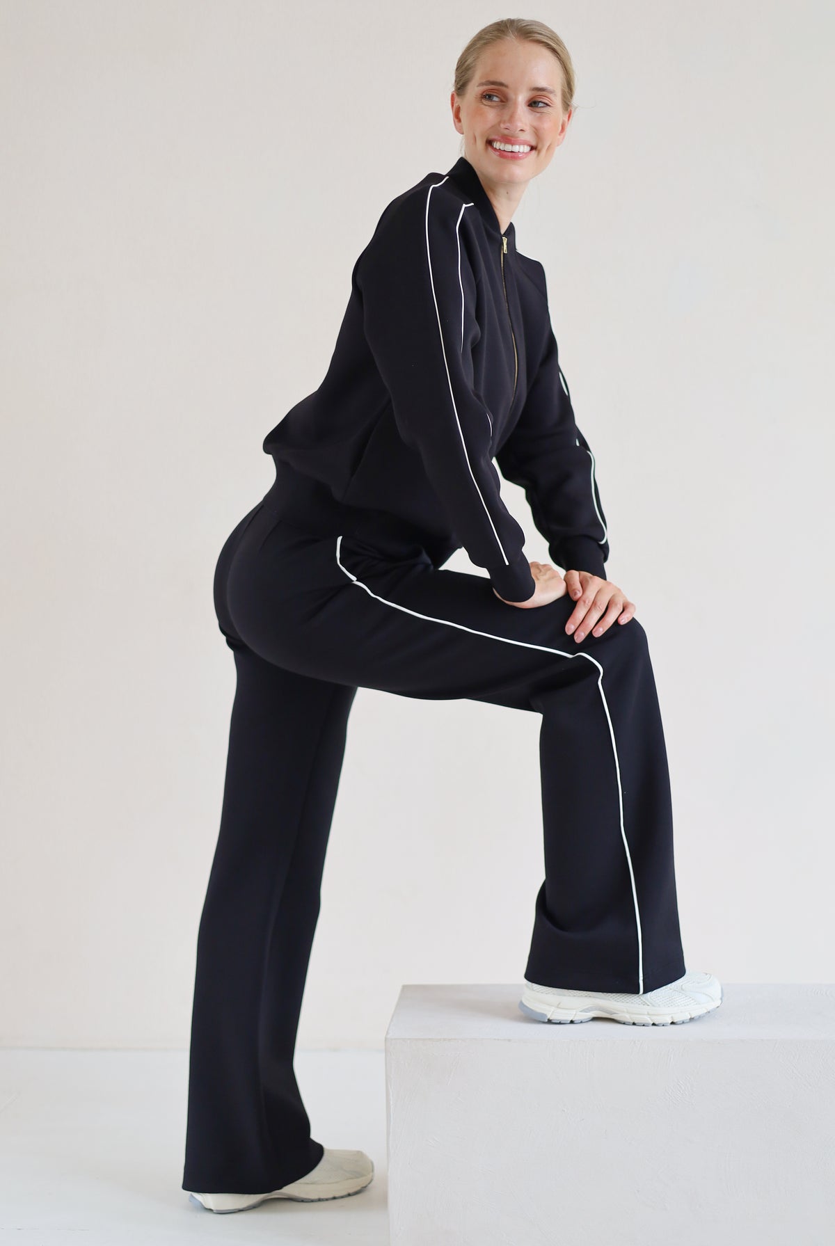A woman modeling a matching black set with white piping details, posing on a white block against a plain white studio background.