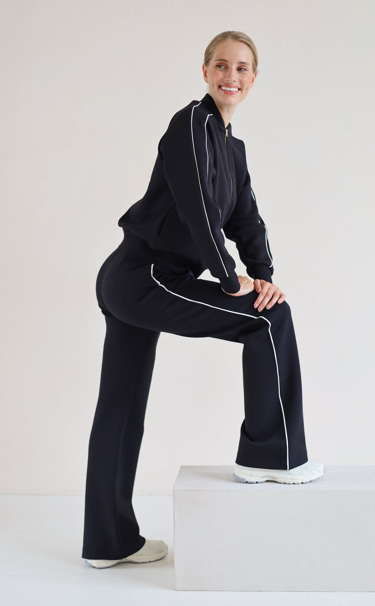 A woman modeling a matching black set with white piping details, posing on a white block against a plain white studio background.
