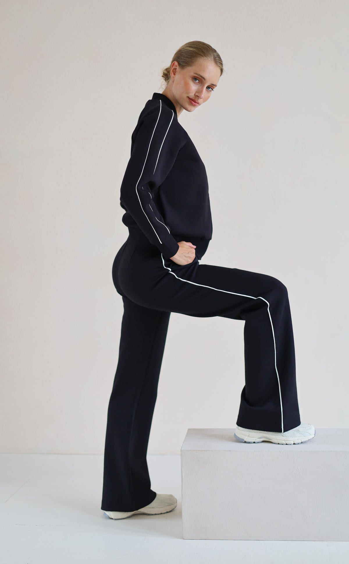 A woman modeling a matching set with white piping details, posing on a white block against a light gray studio background.