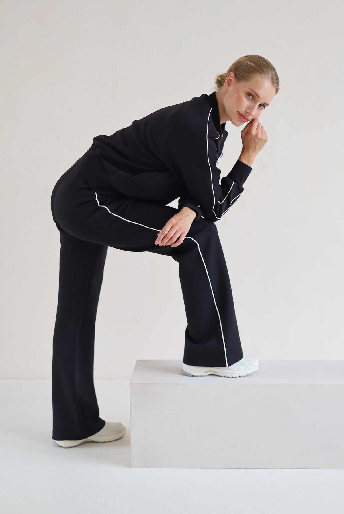 A woman modeling a black athletic set with white piping details, posing on a white platform against a light gray studio background.