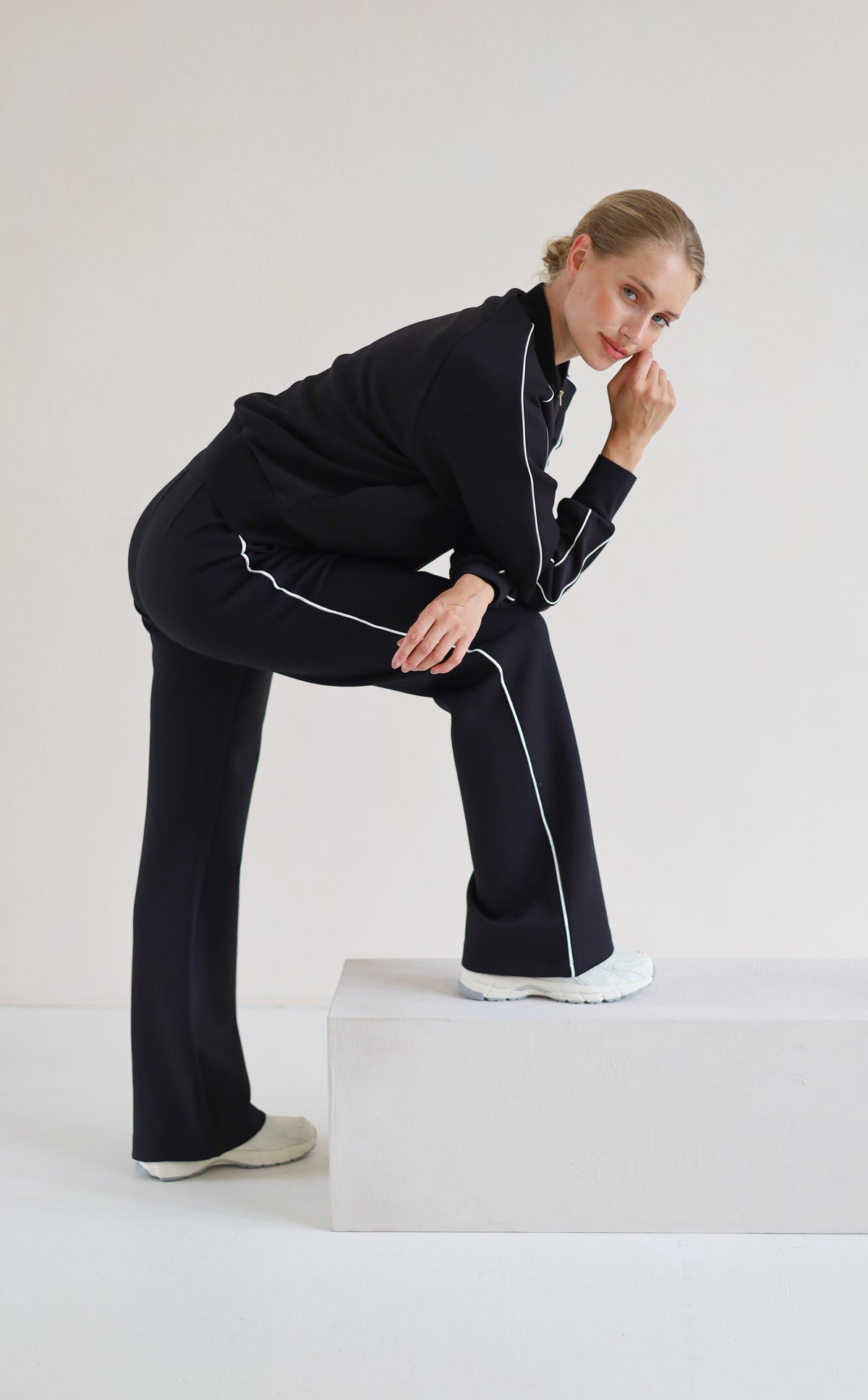 A woman modeling a black athletic set with white piping details, posing on a white platform against a light gray studio background.