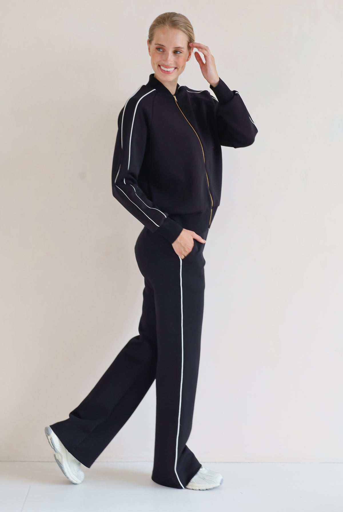 A woman modeling a matching black se with white piping details, featuring a bomber jacket and wide-leg pants, set against a plain light background.