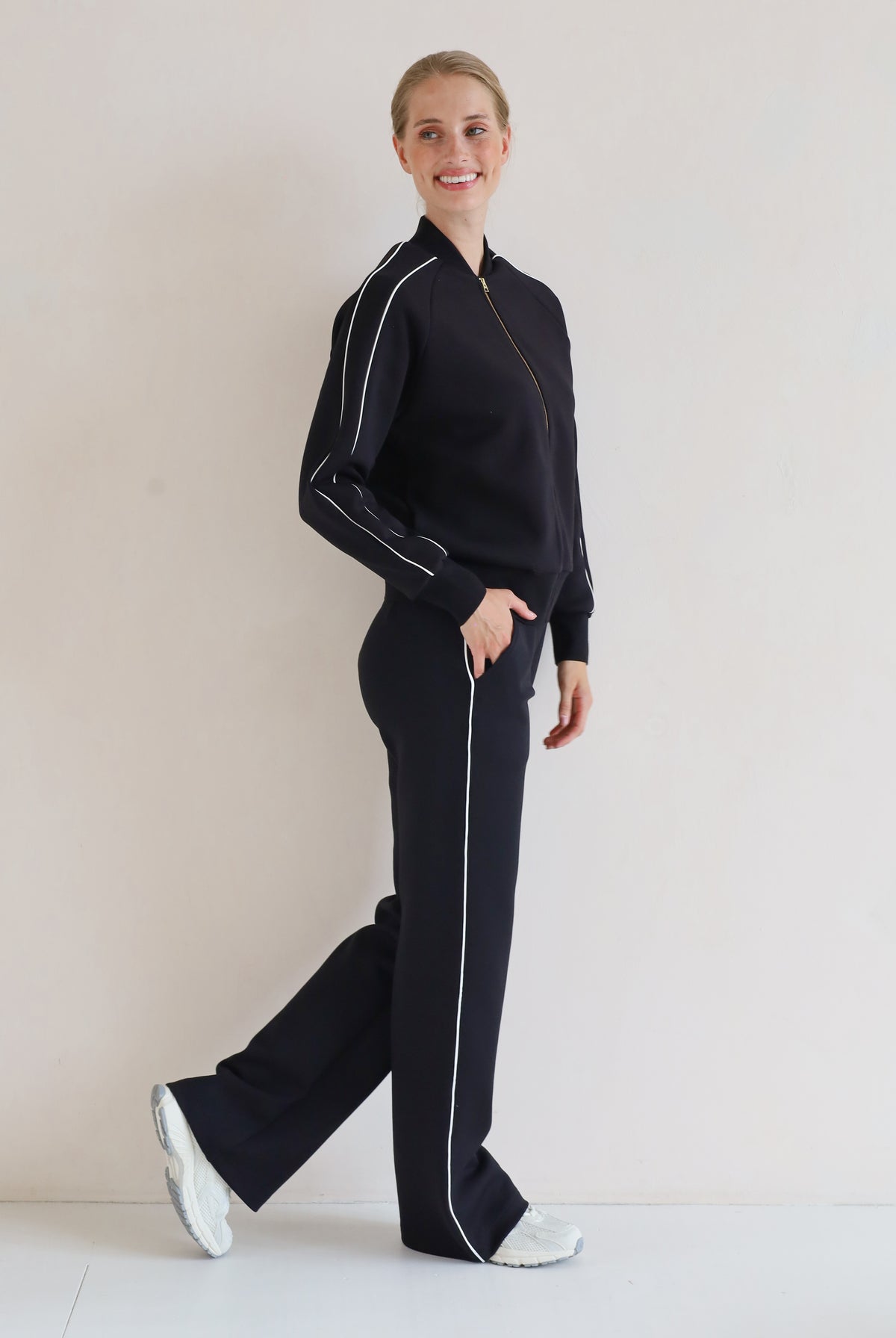 A woman modeling a matching black set with white piping details, consisting of a bomber jacket and wide-leg pants, standing against a plain off-white background.