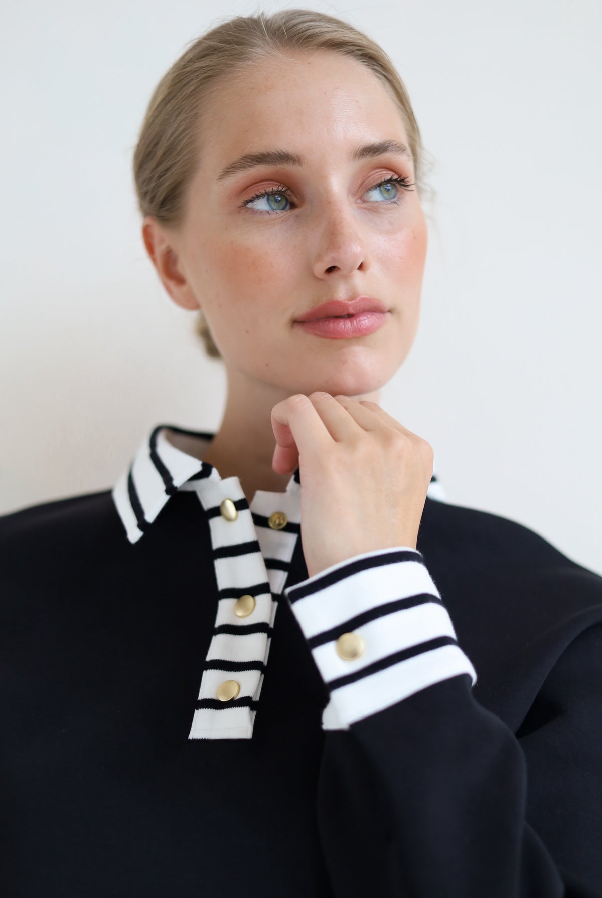 A woman wearing a black knit sweater with a contrasting black and white striped collar and striped cuffs, accented with gold buttons, set against a plain light background.