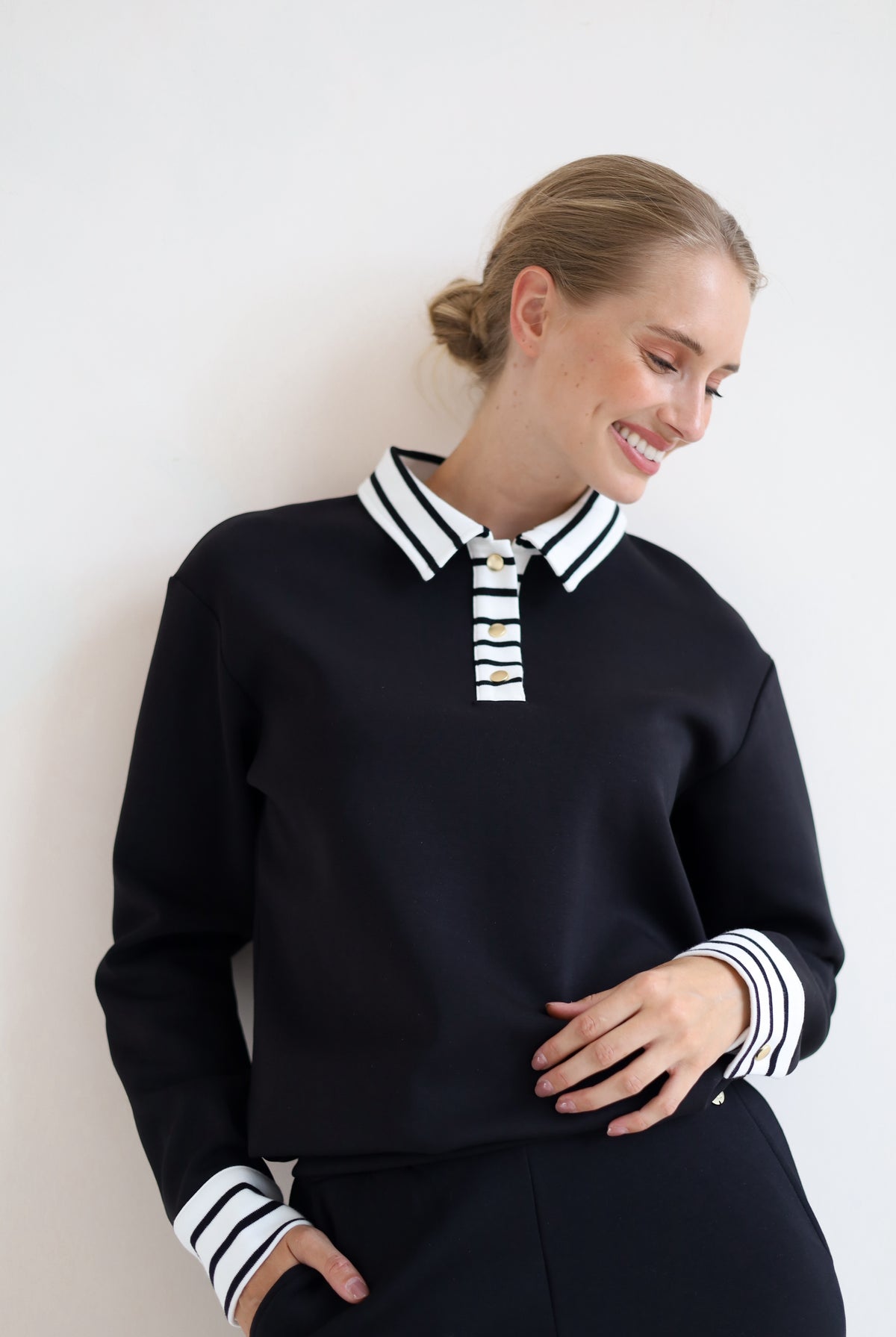 A woman wearing a black long-sleeve knit top with a white and black striped polo-style collar and striped cuffs, paired with black bottoms, posing against a plain white wall.