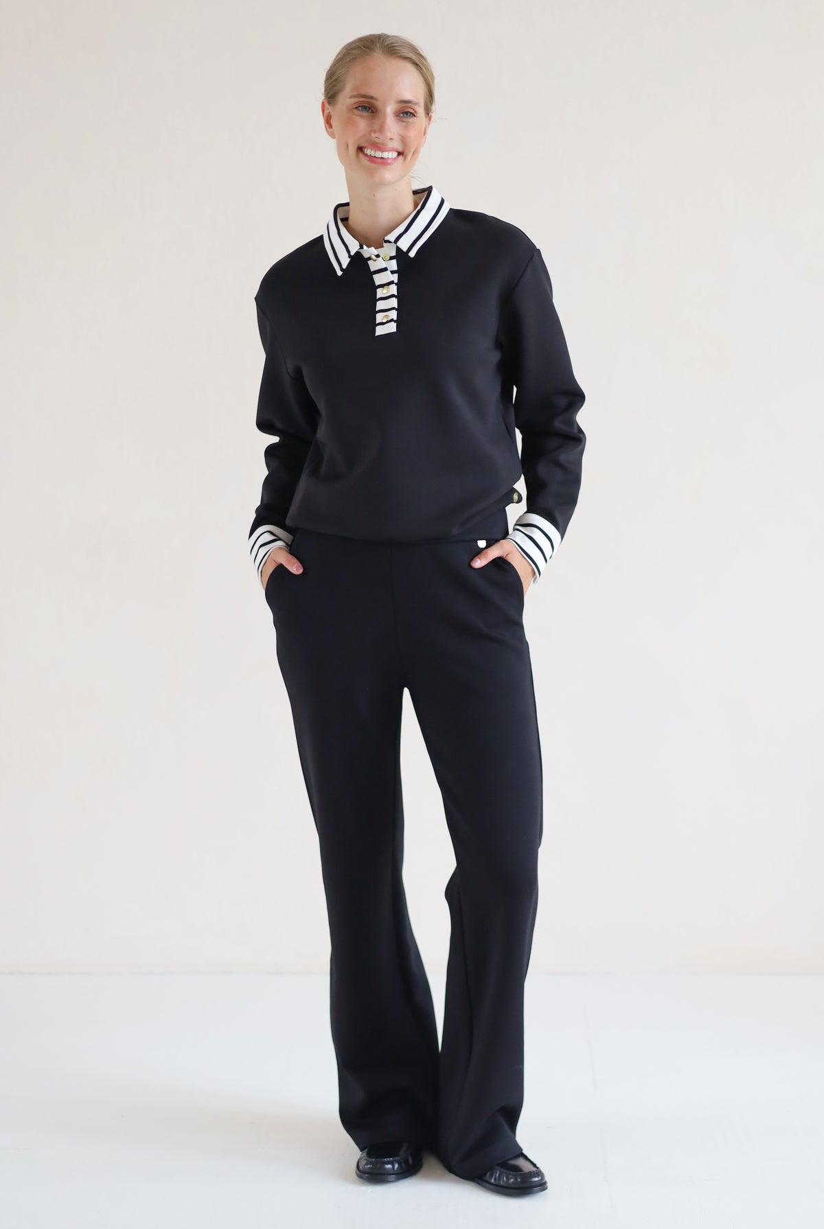 A woman modeling a black long-sleeve polo-style top with a white striped collar and striped cuffs, paired with black wide-leg trousers, against a plain white studio background.