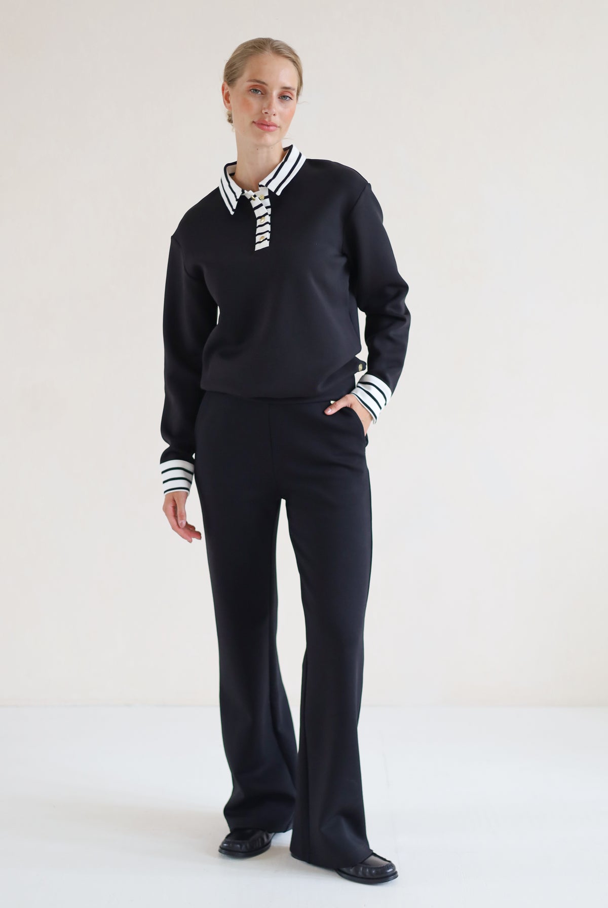 A woman modeling a black knit two-piece set consisting of a long-sleeve polo-style top with striped collar and cuffs, and matching wide-leg trousers, against a plain off-white studio background.
