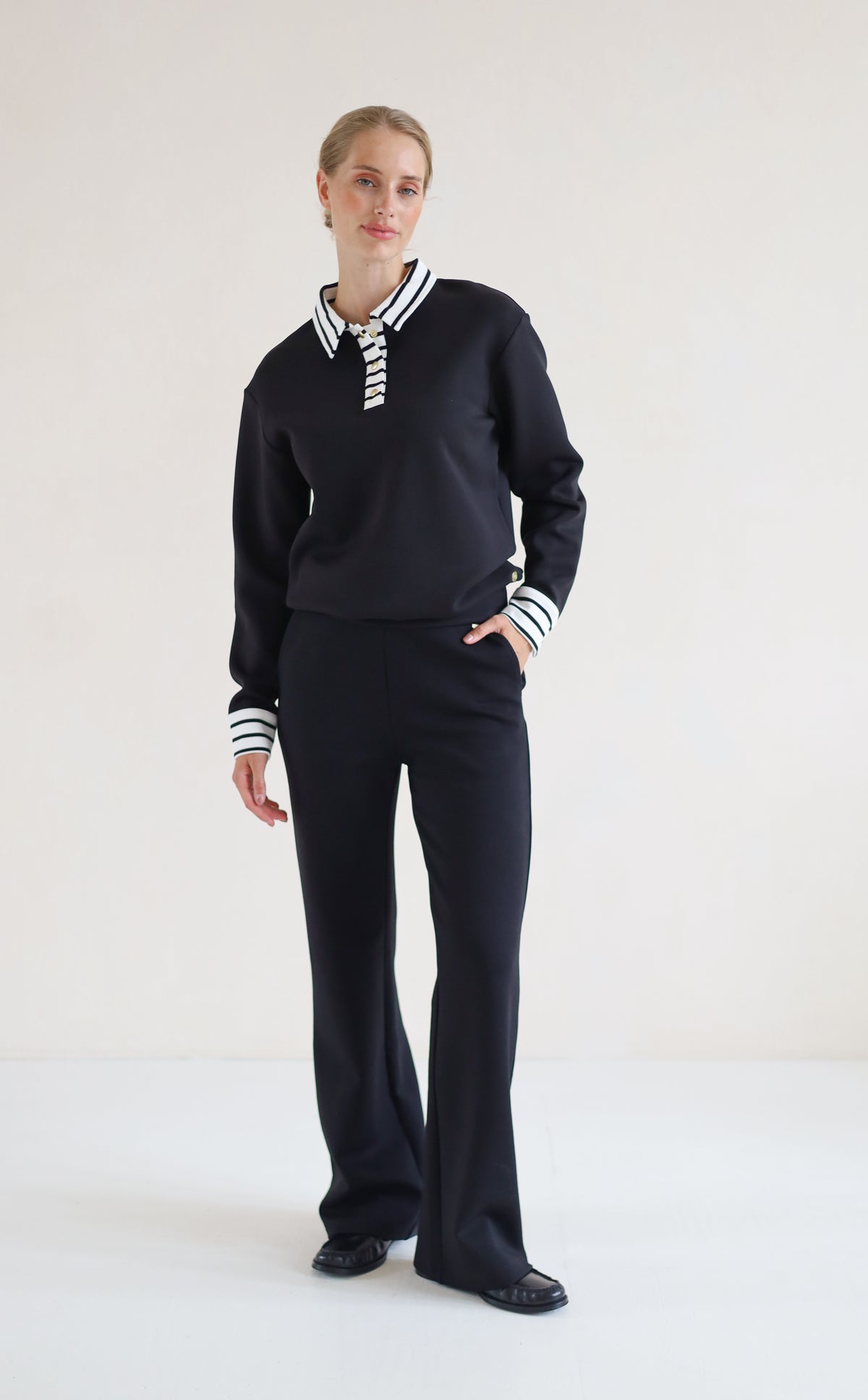 A woman modeling a black knit two-piece set consisting of a long-sleeve polo-style top with striped collar and cuffs, and matching wide-leg trousers, against a plain off-white studio background.