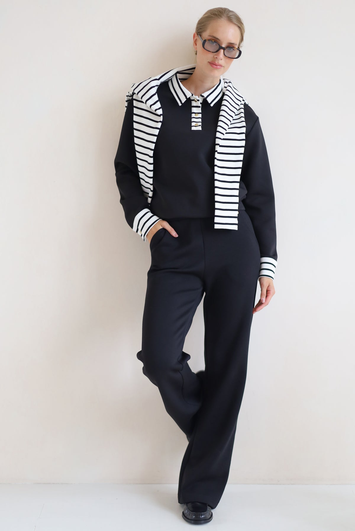 A woman modeling a black knit set consisting of a long-sleeve collared top with black and white horizontal stripes on the shoulders and cuffs, matching black wide-leg trousers, and black shoes, set against a plain off-white wall.
