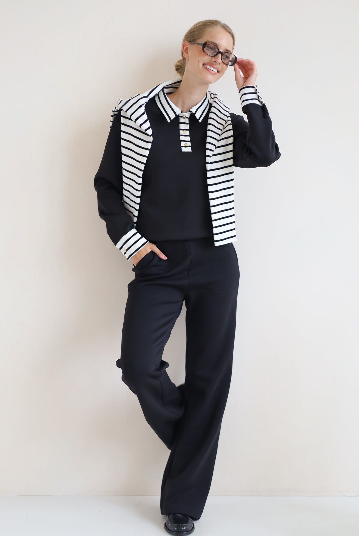 A woman modeling a black knit set consisting of a long-sleeve collared top with black and white horizontal stripes on the shoulders and cuffs, matching black wide-leg trousers, and black shoes, set against a plain off-white wall.
