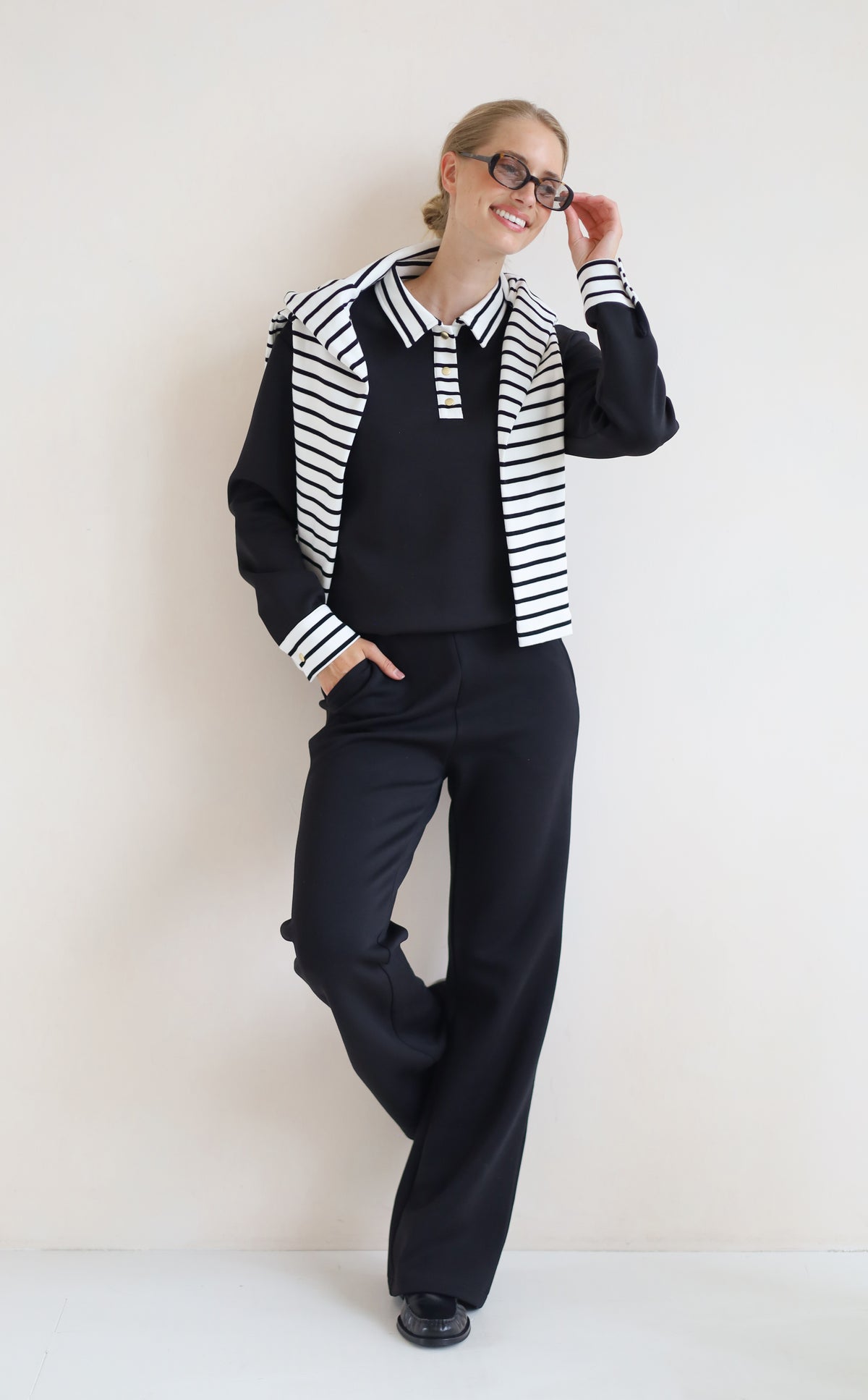 A woman modeling a black knit set consisting of a long-sleeve collared top with black and white horizontal stripes on the shoulders and cuffs, matching black wide-leg trousers, and black shoes, set against a plain off-white wall.
