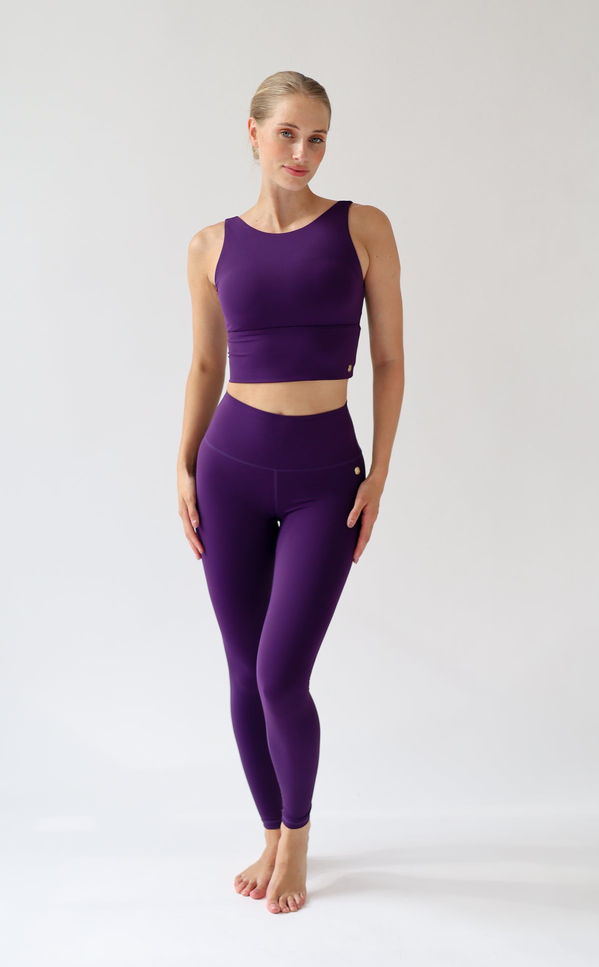 A woman modeling a matching purple activewear set consisting of a sleeveless crop top and high-waisted leggings, standing against a plain light gray studio background.