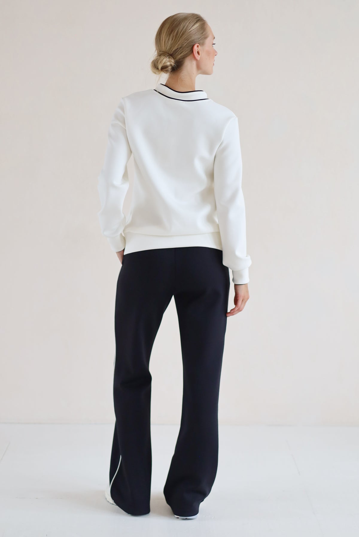 A woman is shown from the back wearing a white v-neck sweater with black trim and black wide-leg trousers, standing against a plain light beige studio background.