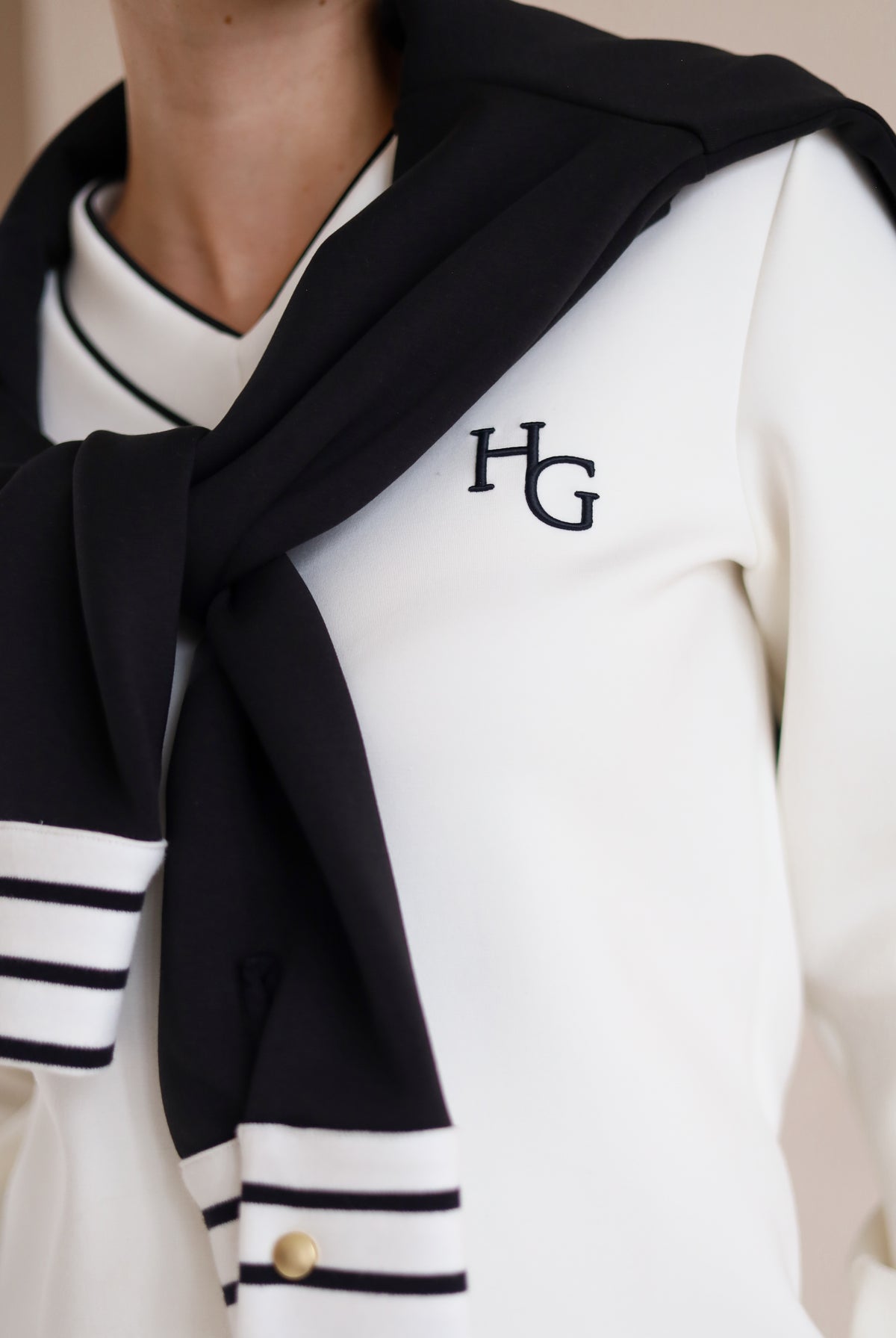 House of Gravity V-Neck Sweater in Off-White Marble and Black, close-up of the embroidered HG logo, styled with the Button Polo Sweater in Black draped over the shoulders.