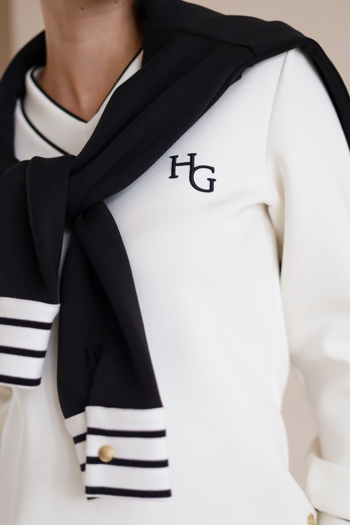 House of Gravity V-Neck Sweater in Off-White Marble and Black, close-up of the embroidered HG logo, styled with the Button Polo Sweater in Black draped over the shoulders.