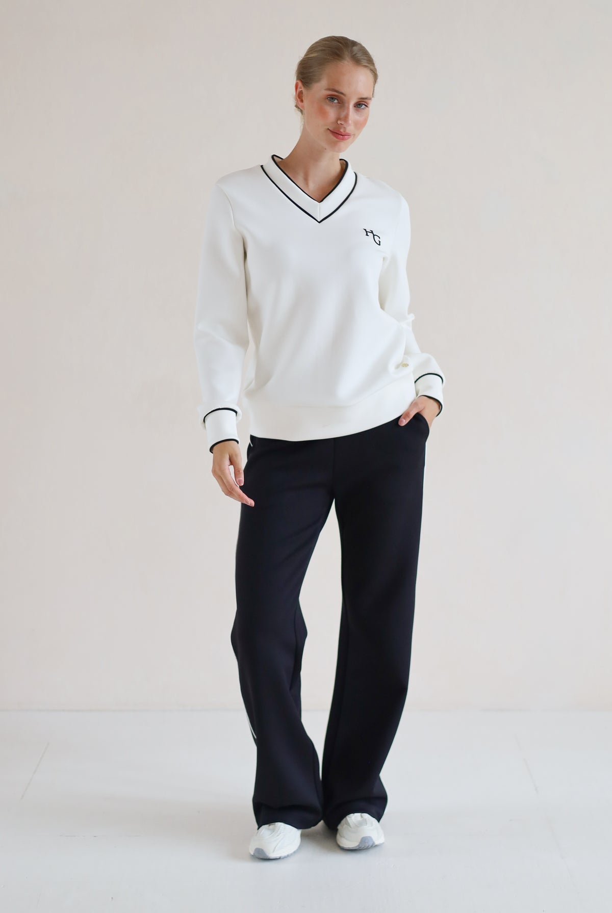 A woman modeling a white long-sleeve V-neck sweater with black trim and a small embroidered chest logo, paired with black wide-leg pants and white sneakers, against a plain light beige studio background.