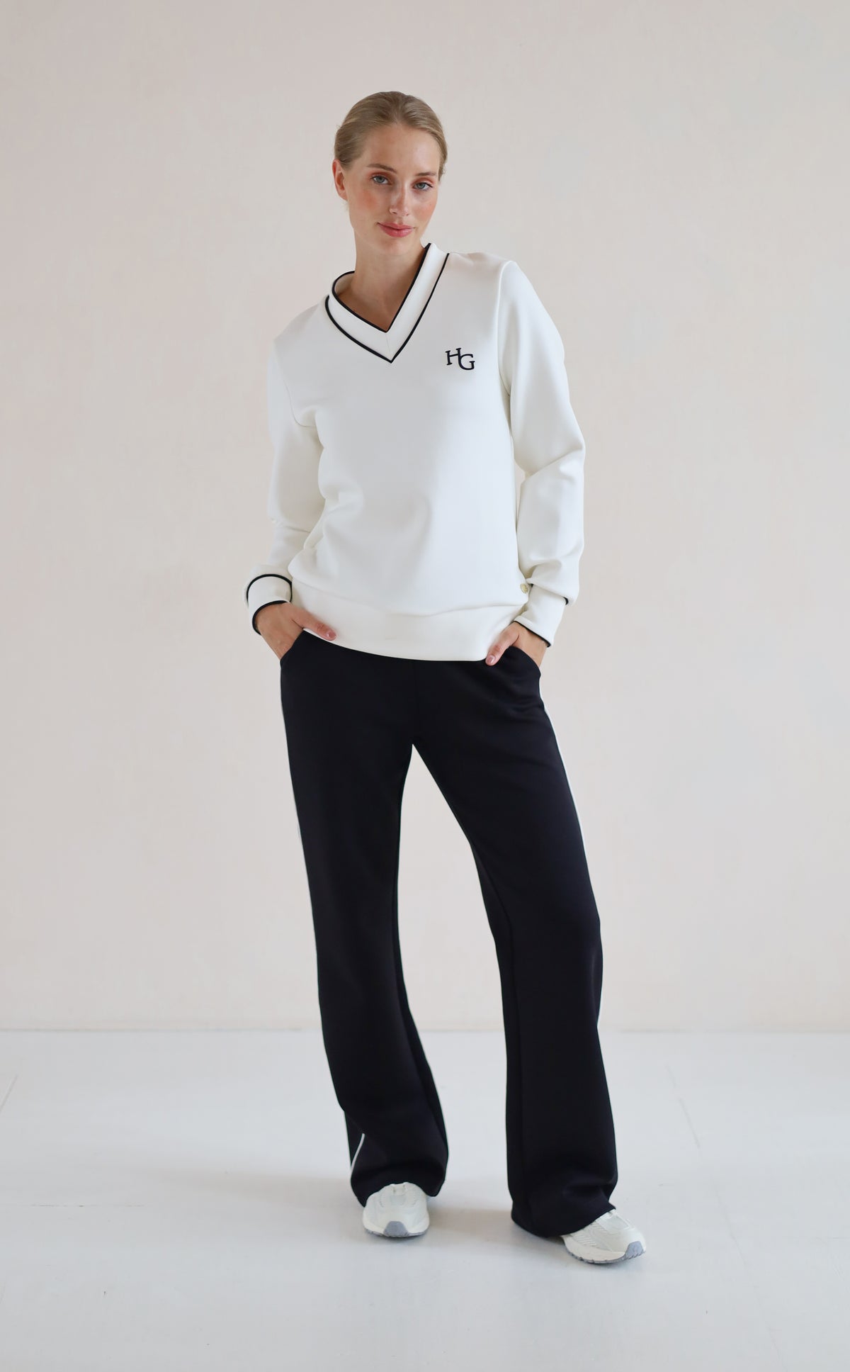 A woman modeling a white long-sleeve V-neck sweatshirt with black trim and a small black 'hg' logo on the chest, paired with black wide-leg sweatpants and white sneakers, set against a plain off-white studio background.