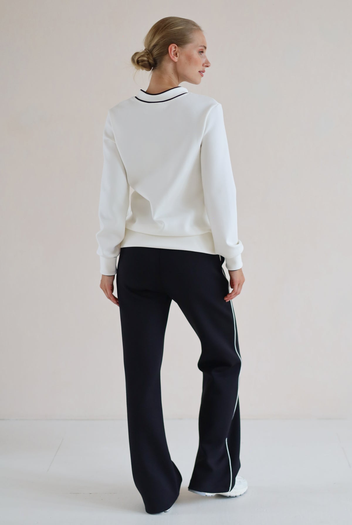 A woman shown from the back wearing a white long-sleeve sweatshirt with a black and white striped collar, paired with black wide-leg trouser featuring thin white side stripes, set against a plain light beige studio background.