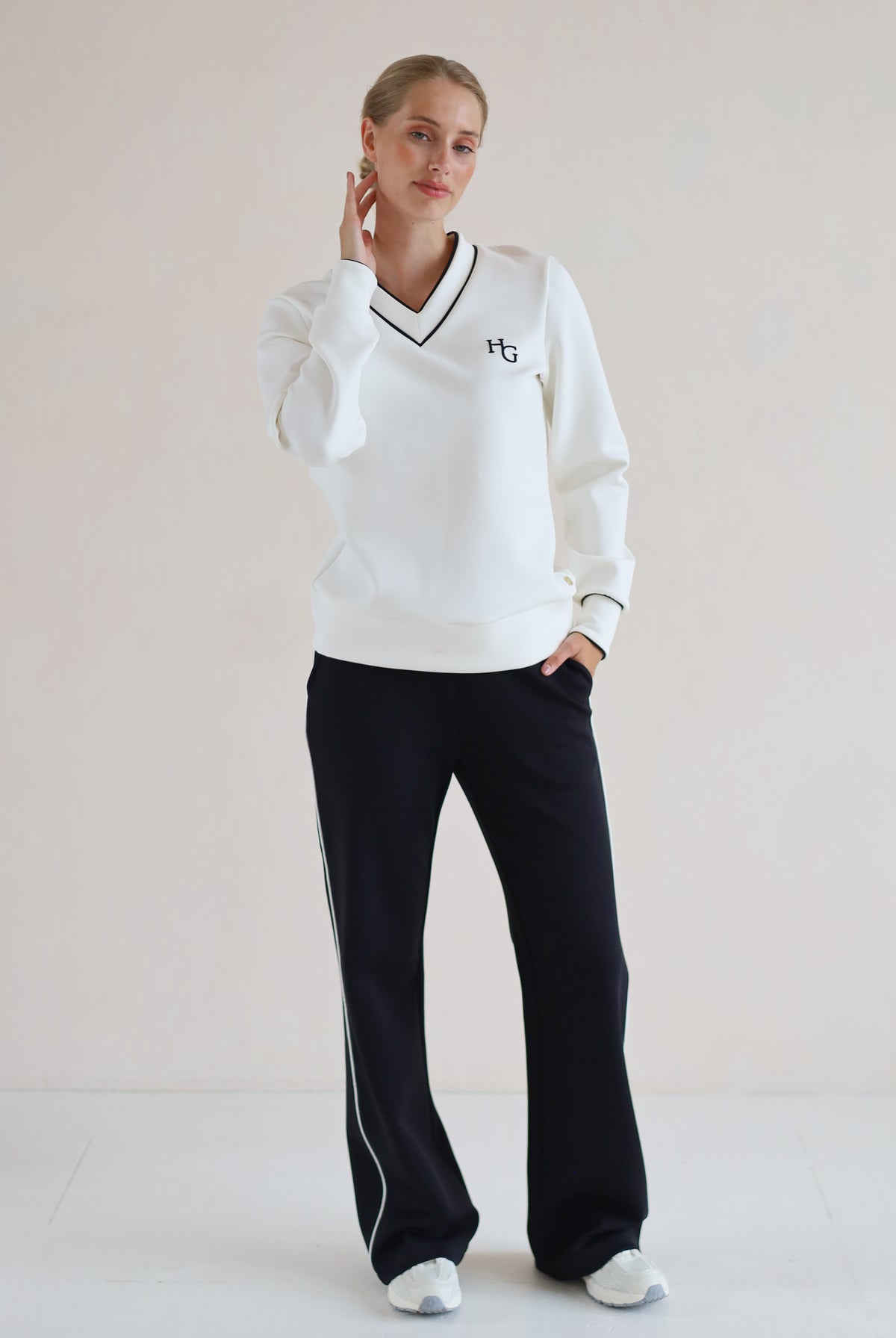 A woman modeling a white long-sleeve V-neck sweatshirt with black trim and a small black monogram logo on the left chest, paired with black wide-leg trouser featuring white side stripes, against a plain light beige studio background.