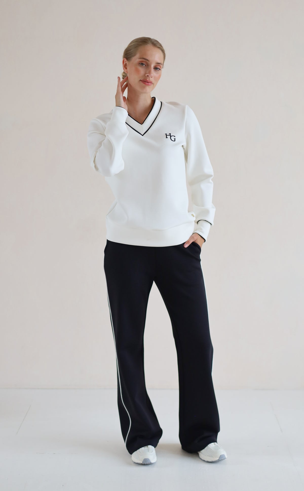 A woman modeling a white long-sleeve V-neck sweatshirt with black trim and a small black monogram logo on the left chest, paired with black wide-leg trouser featuring white side stripes, against a plain light beige studio background.