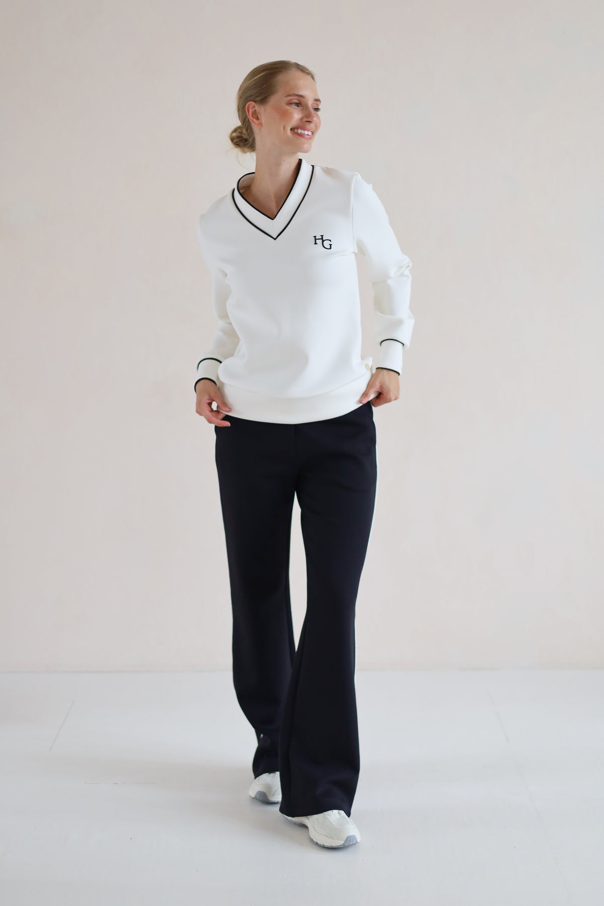 A woman modeling a white long-sleeve V-neck sweater with black trim and a small embroidered logo on the chest, paired with black pants and white sneakers, in a minimalist studio setting.