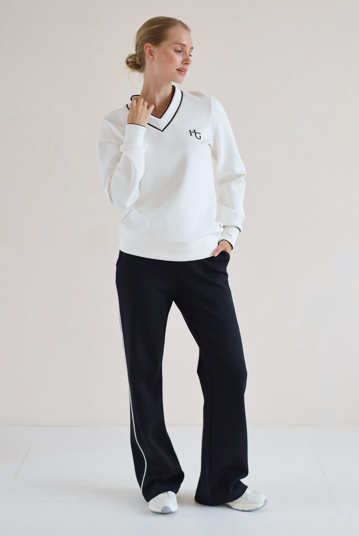 A woman modeling a white long-sleeve V-neck sweatshirt with black trim and a small embroidered logo on the chest, paired with black wide-leg trouser and white sneakers, against a plain light beige studio background.