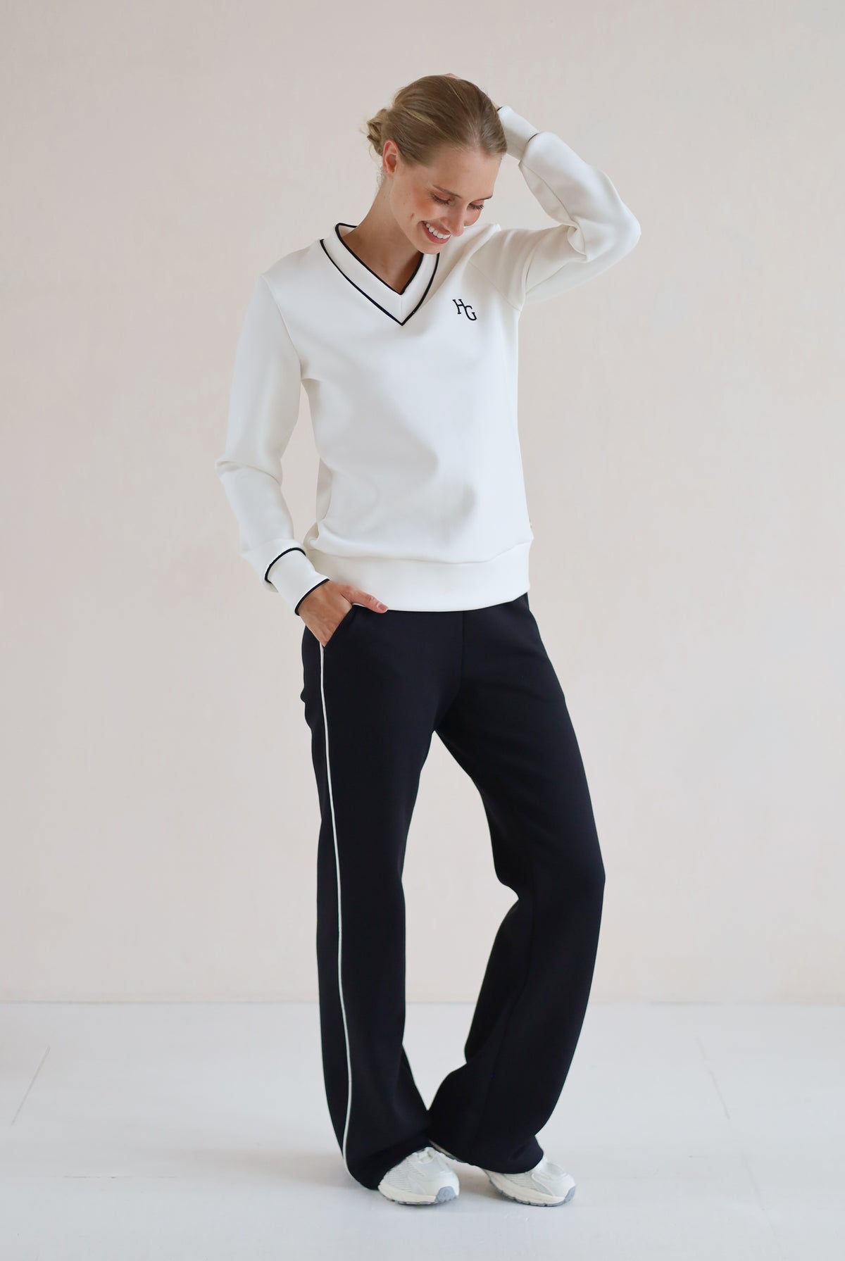 A woman modeling a white long-sleeve V-neck sweatshirt with black trim and a small embroidered logo on the chest, paired with black trouser featuring a white side stripe, against a plain light beige studio background.