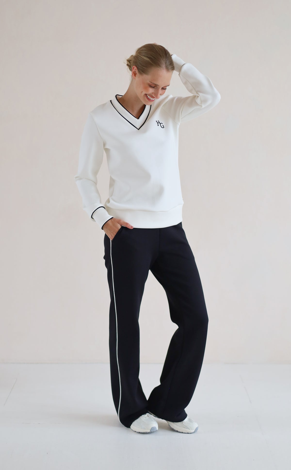 A woman modeling a white long-sleeve V-neck sweatshirt with black trim and a small embroidered logo on the chest, paired with black trouser featuring a white side stripe, against a plain light beige studio background.