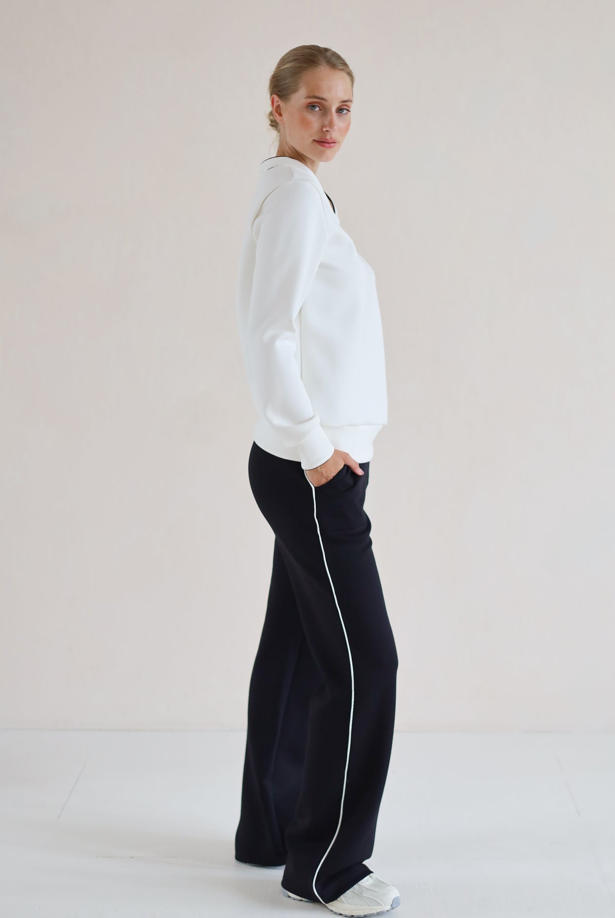 A woman stands in profile wearing a white long-sleeve V-Neck Sweater and black wide-leg pants with a white side stripe, against a plain light beige studio background.