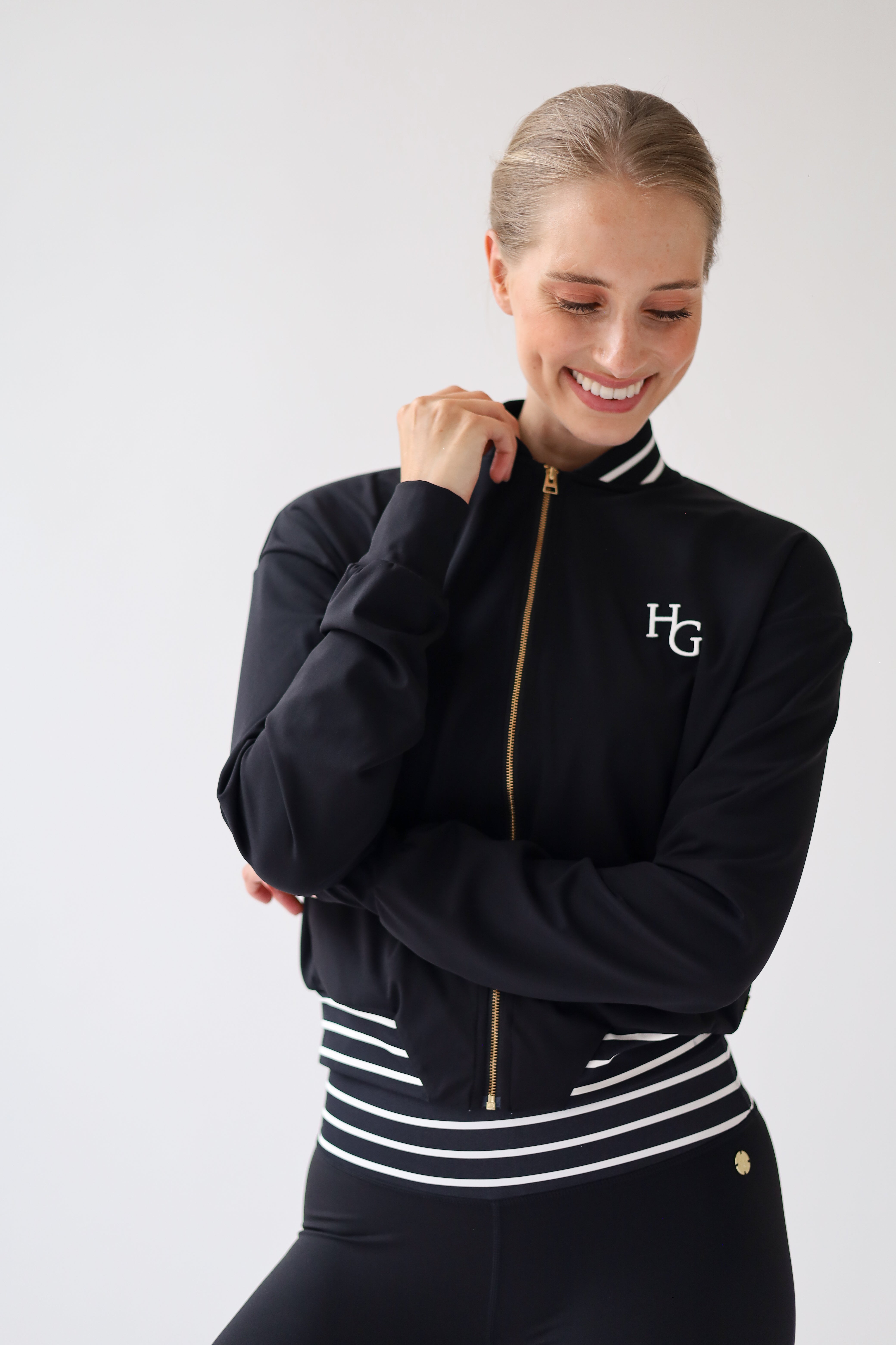 A woman wearing a black zip-up athletic jacket with white stripes on the collar and hem, gold zipper details, and a white 'HG' logo on the left chest, posed against a light gray studio background.