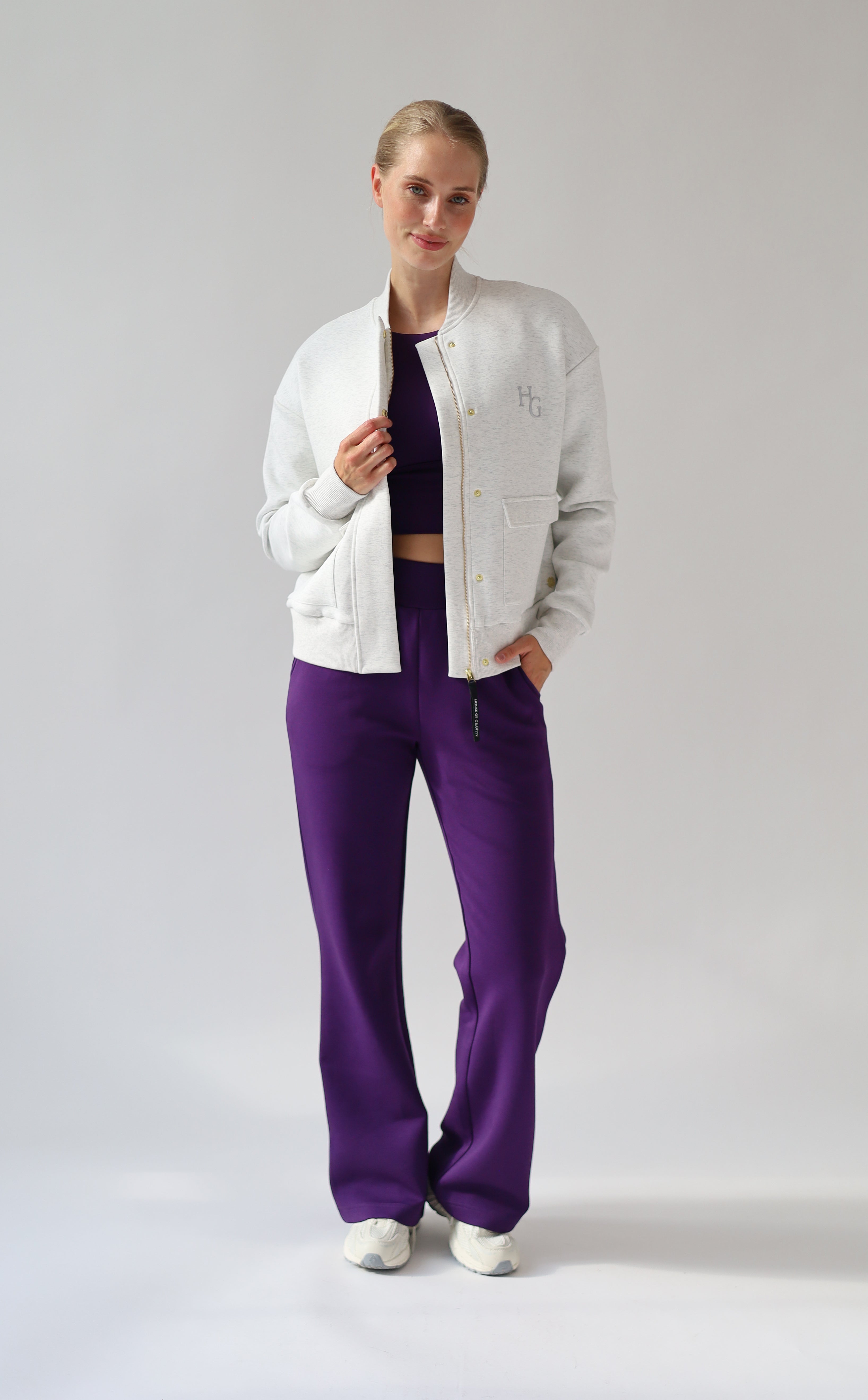 A model posing in a studio setting wearing a light grey melange bomber jacket, a dark purple crop top, and vibrant purple wide-leg trousers.