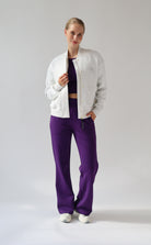 A model posing in a studio setting wearing a light grey melange bomber jacket, a dark purple crop top, and vibrant purple wide-leg trousers.