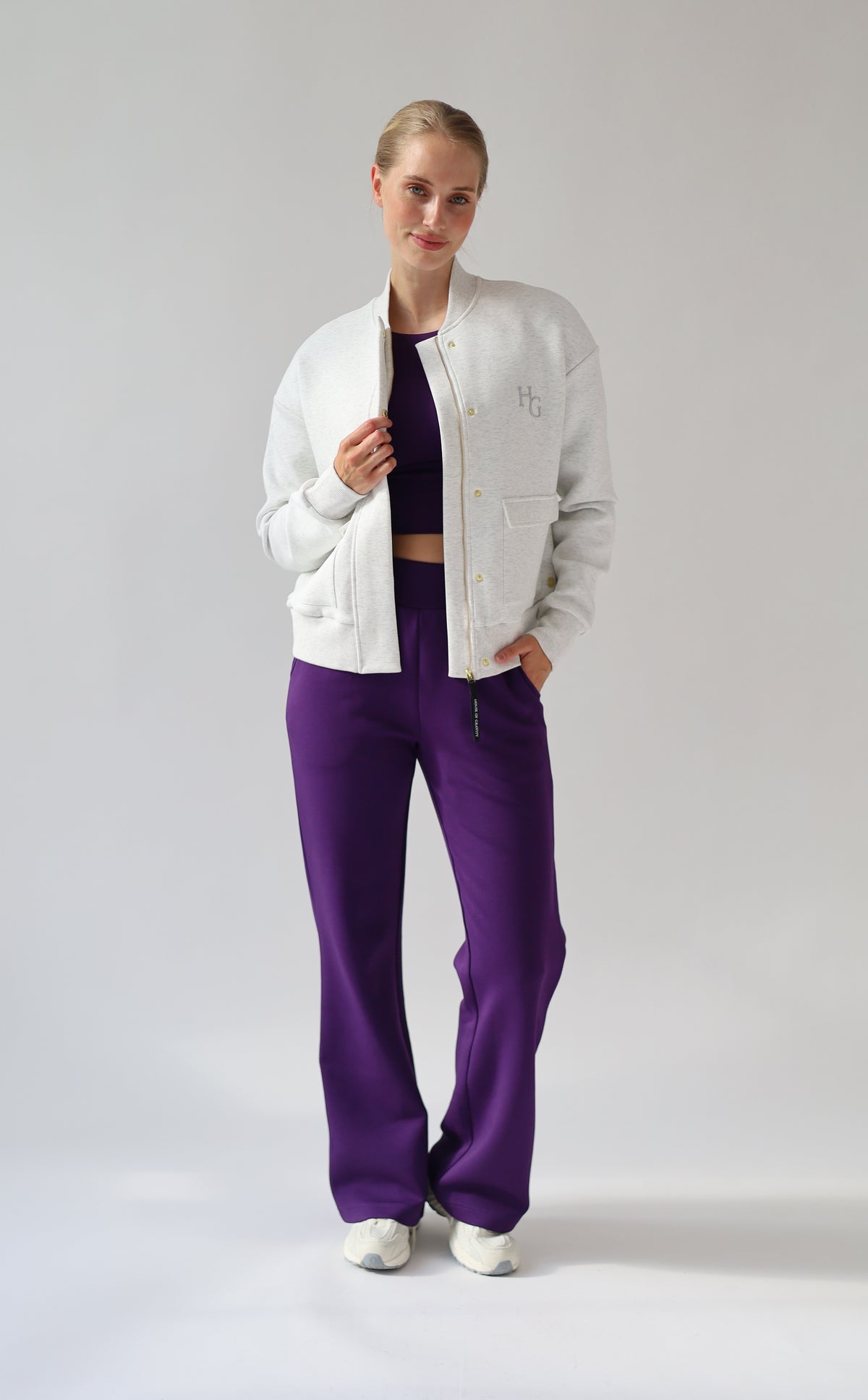 A model posing in a studio setting wearing a light grey melange bomber jacket, a dark purple crop top, and vibrant purple wide-leg trousers.