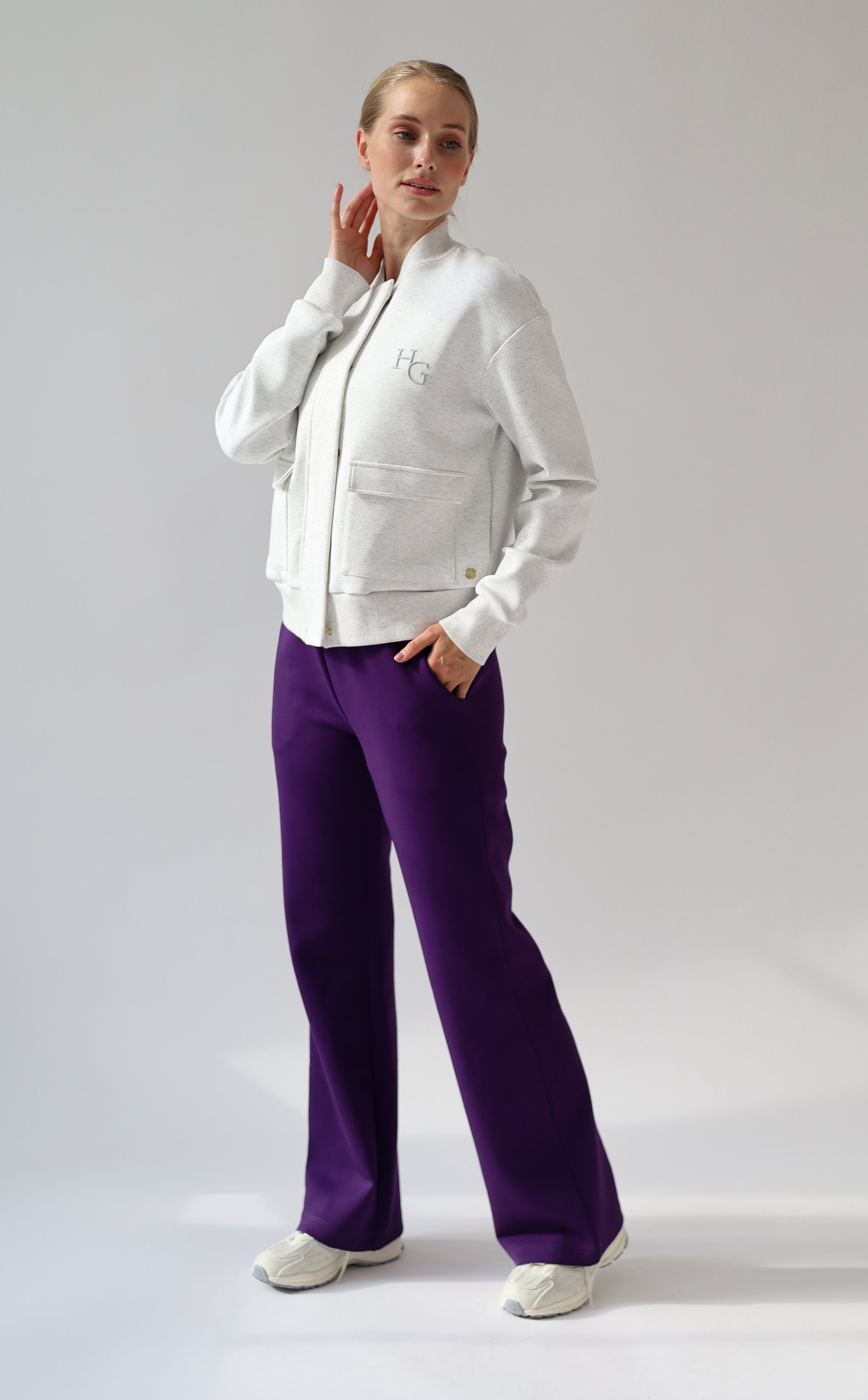 A model posing in a studio setting wearing a light grey melange bomber jacket, a dark purple crop top, and vibrant purple wide-leg trousers.