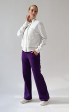 A model posing in a studio setting wearing a light grey melange bomber jacket, a dark purple crop top, and vibrant purple wide-leg trousers.