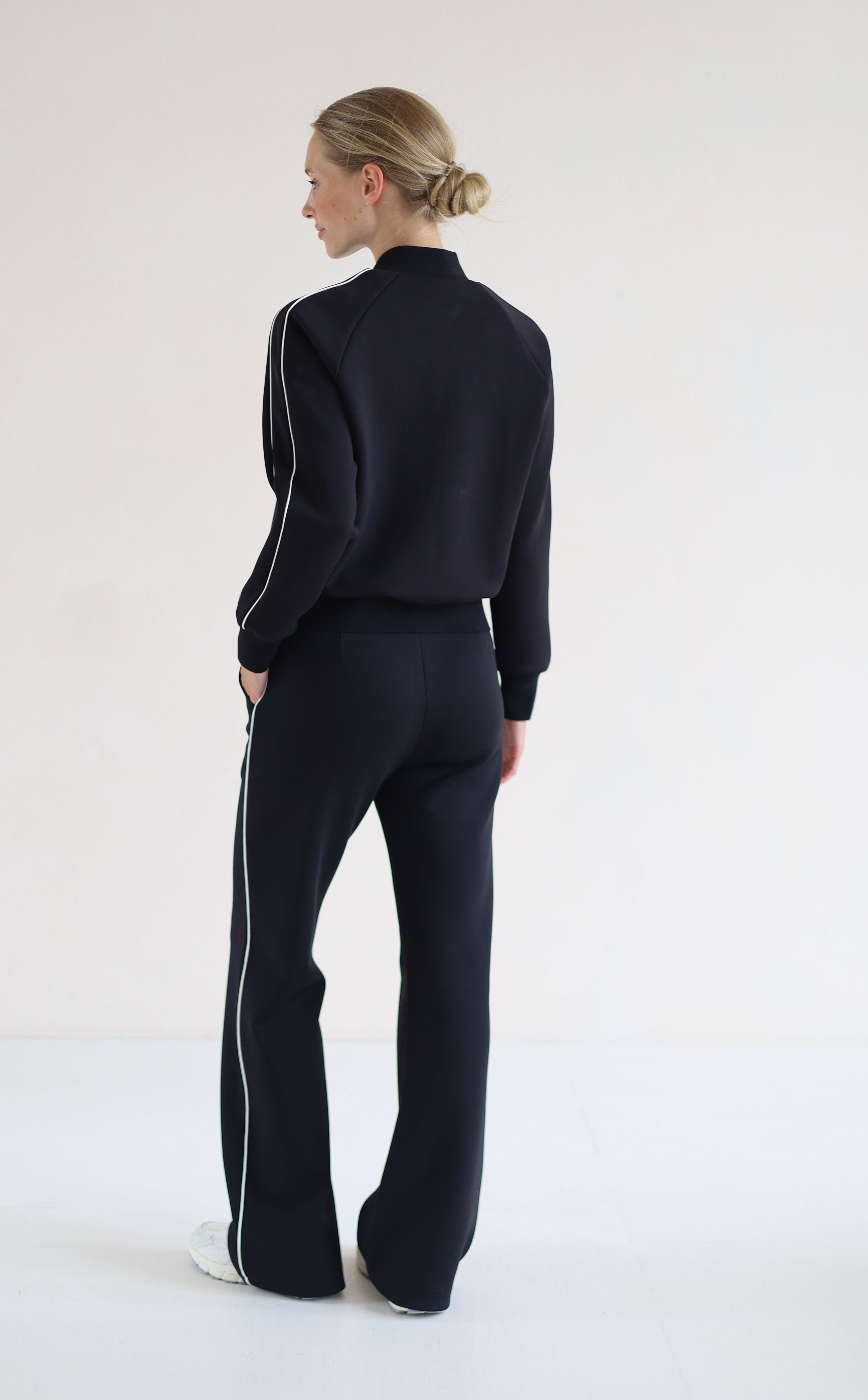 A woman stands with her back to the camera wearing a matching black set with white side stripes, consisting of a cropped mock-neck jacket and wide-leg pants, against a plain white studio background.