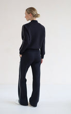 A woman stands with her back to the camera wearing a matching black set with white side stripes, consisting of a cropped mock-neck jacket and wide-leg pants, against a plain white studio background.