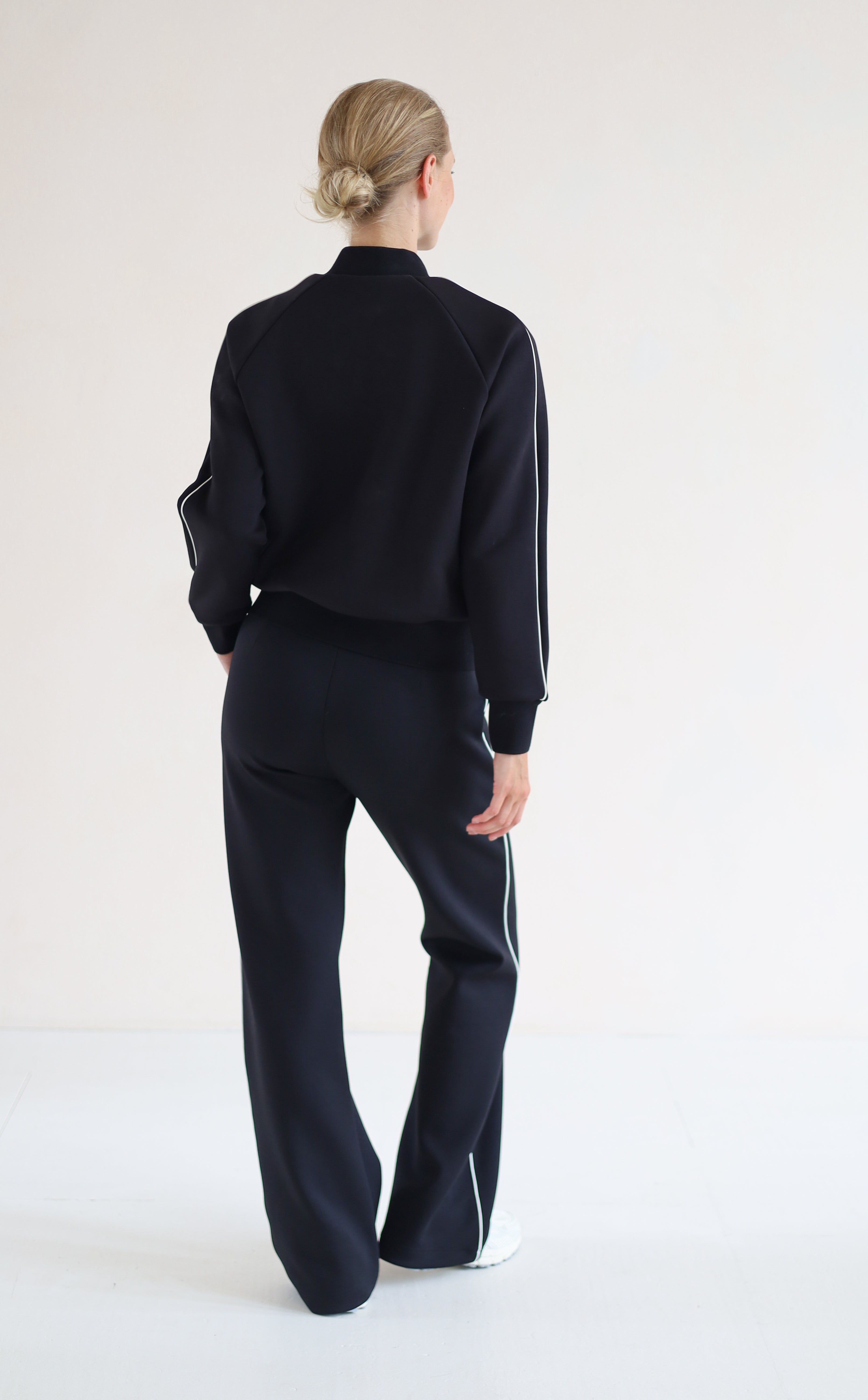 A woman viewed from behind wearing a blackbomber jacket with white side stripes and matching black wide-leg trousers with white side stripes, standing against a plain white studio background.