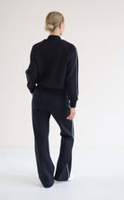A woman viewed from behind wearing a blackbomber jacket with white side stripes and matching black wide-leg trousers with white side stripes, standing against a plain white studio background.